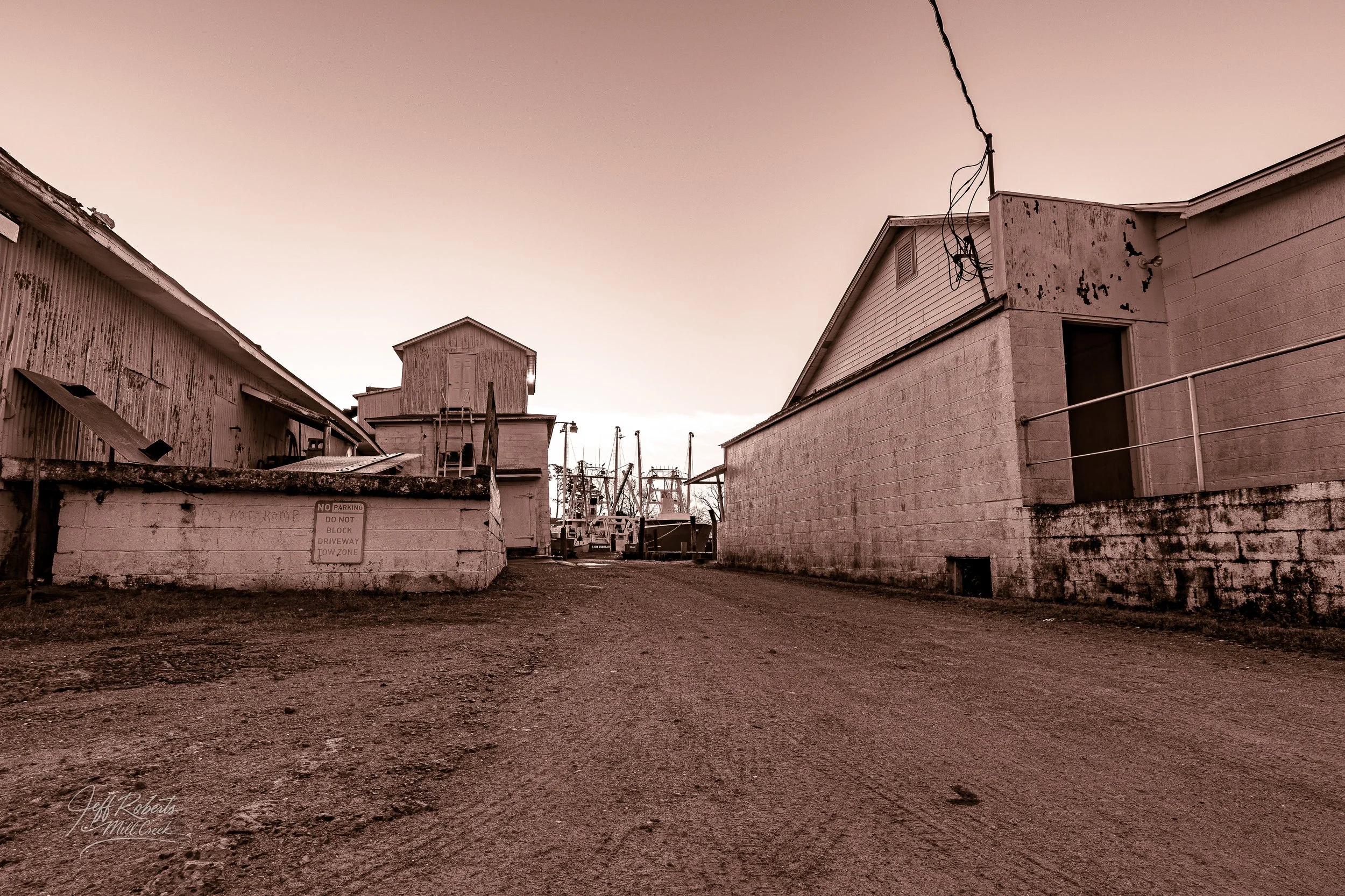 A dirt alleyway between weathered buildings with boats and masts in the distance, under a pinkish sky.