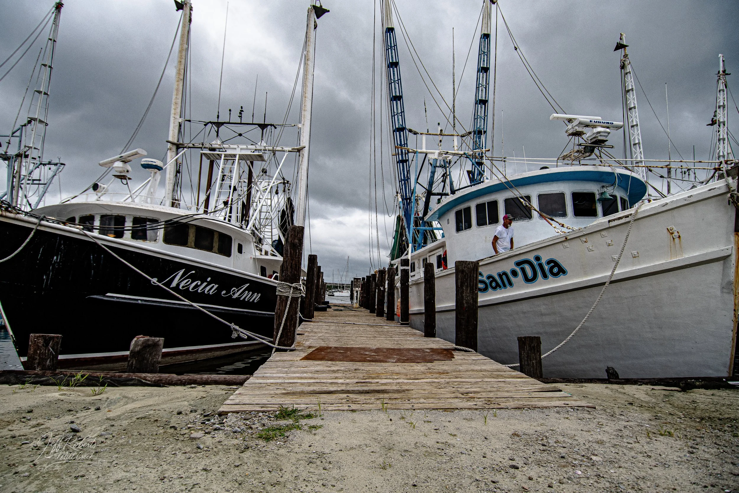 View of boats docked at a marina with a wooden pier in the foreground and dark, cloudy sky overhead.