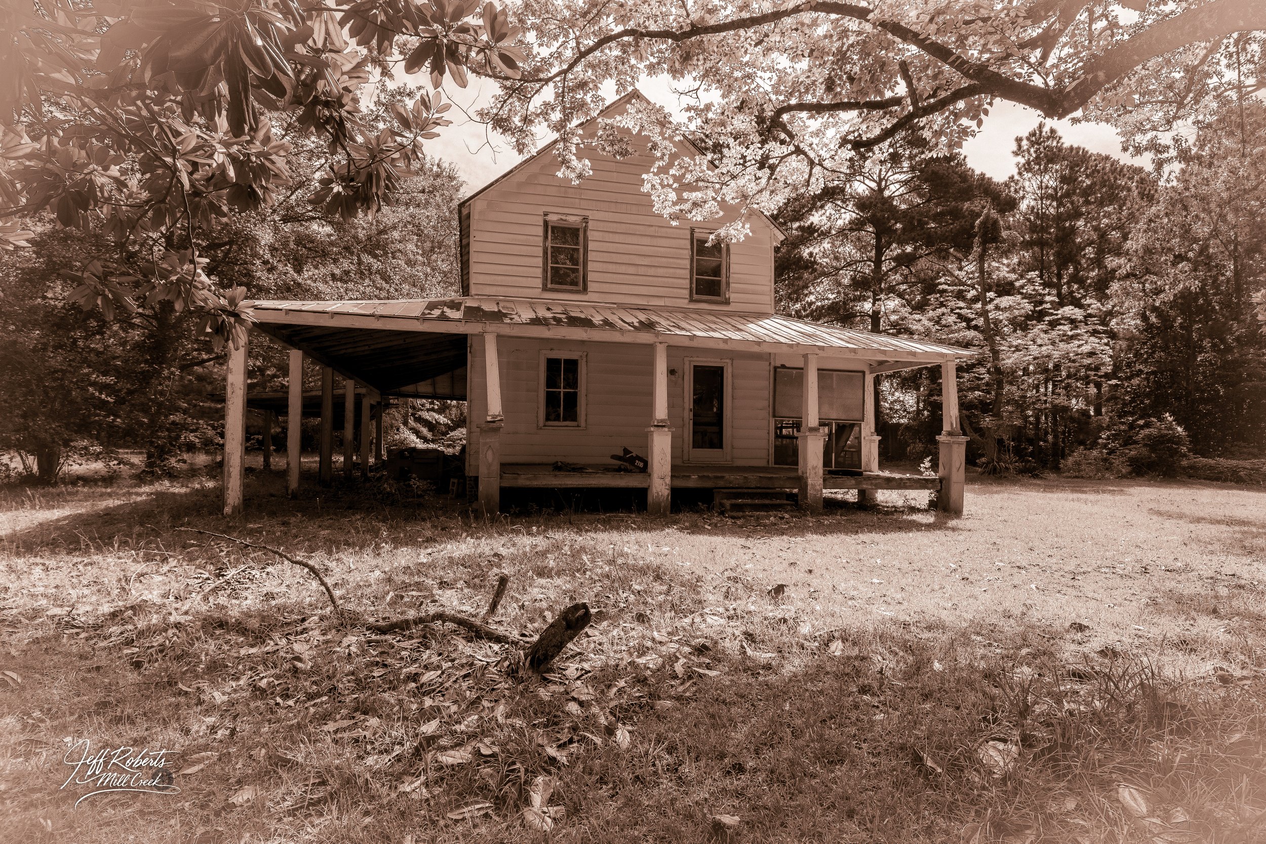Sepia-toned photo of an old, two-story house with a large porch. The house has peeling paint, overgrown grass, and is surrounded by trees. The scene appears to be in rural or wooded area.