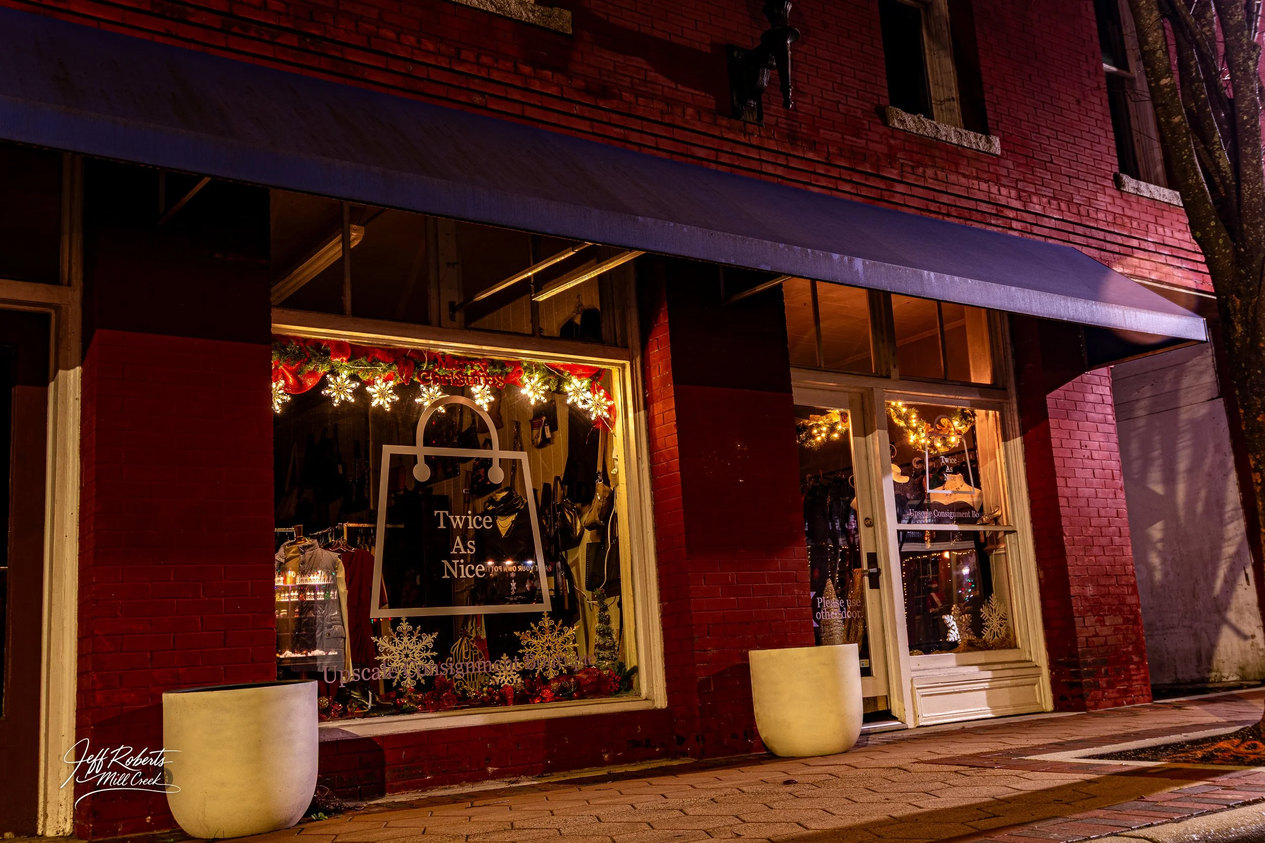 The storefront of a retail shop decorated for Christmas with illuminated snowflakes and garlands, with a sign reading 'Twice As Nice' displayed in the window.