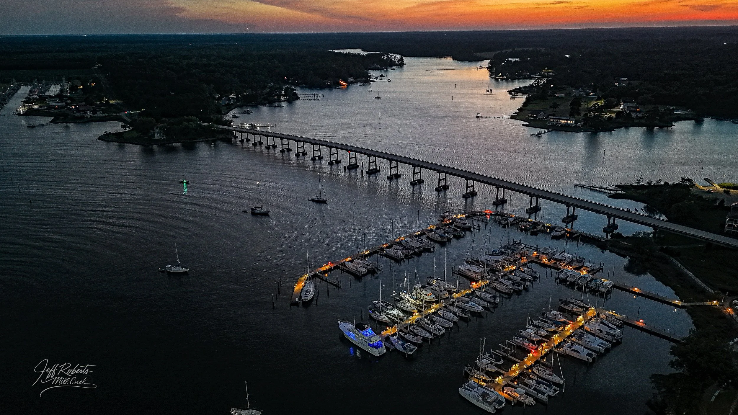 Aerial view of a marina with boats docked at night, illuminated by streetlights, with a bridge crossing over the water, houses on the shoreline, and a sunset sky in the background.