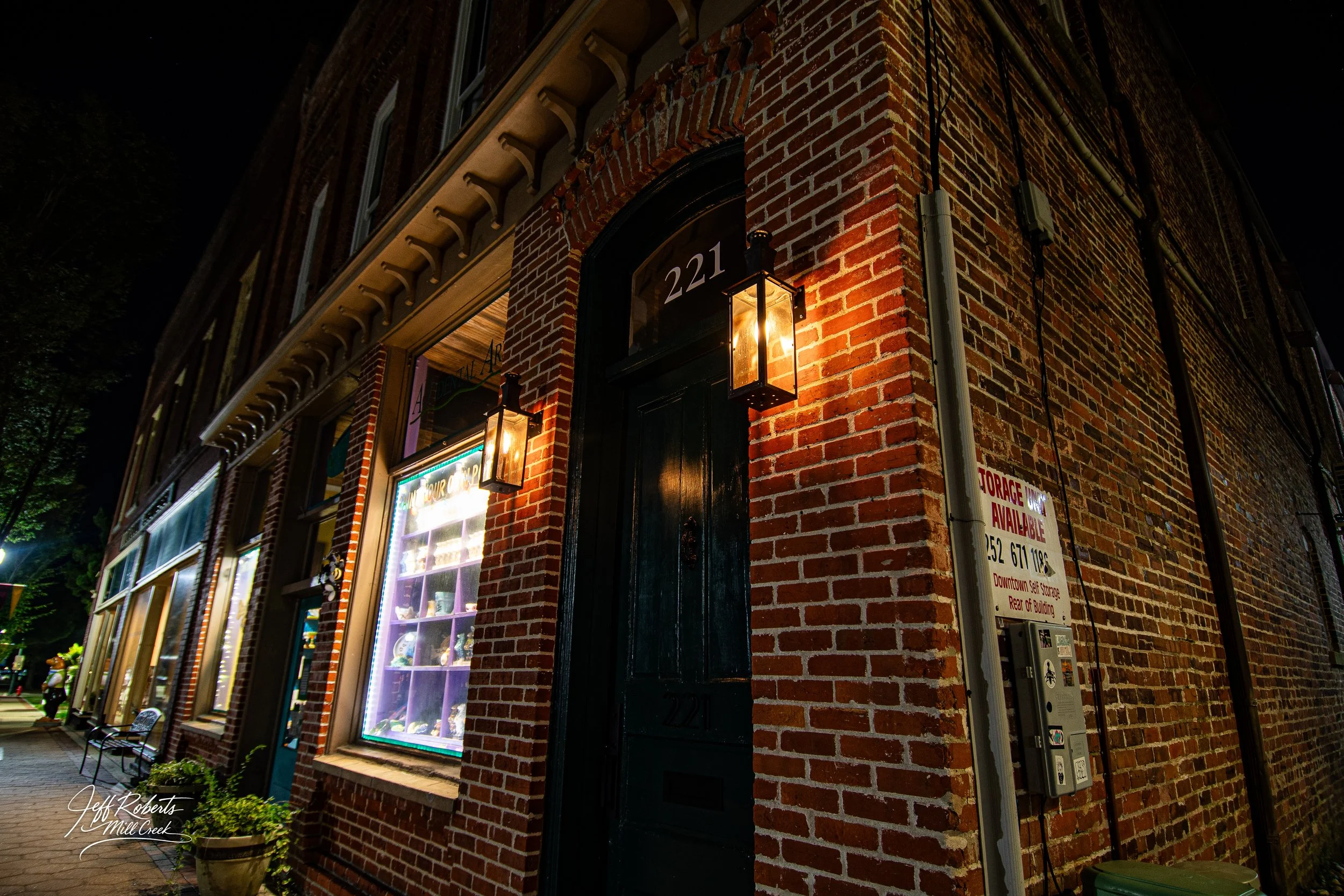 Nighttime exterior of a brick building with two lit lanterns flanking a black door, numbered 221. The door and windows are part of a storefront, with a sign indicating storage availability on the side. A sidewalk with potted plants and a bench runs a