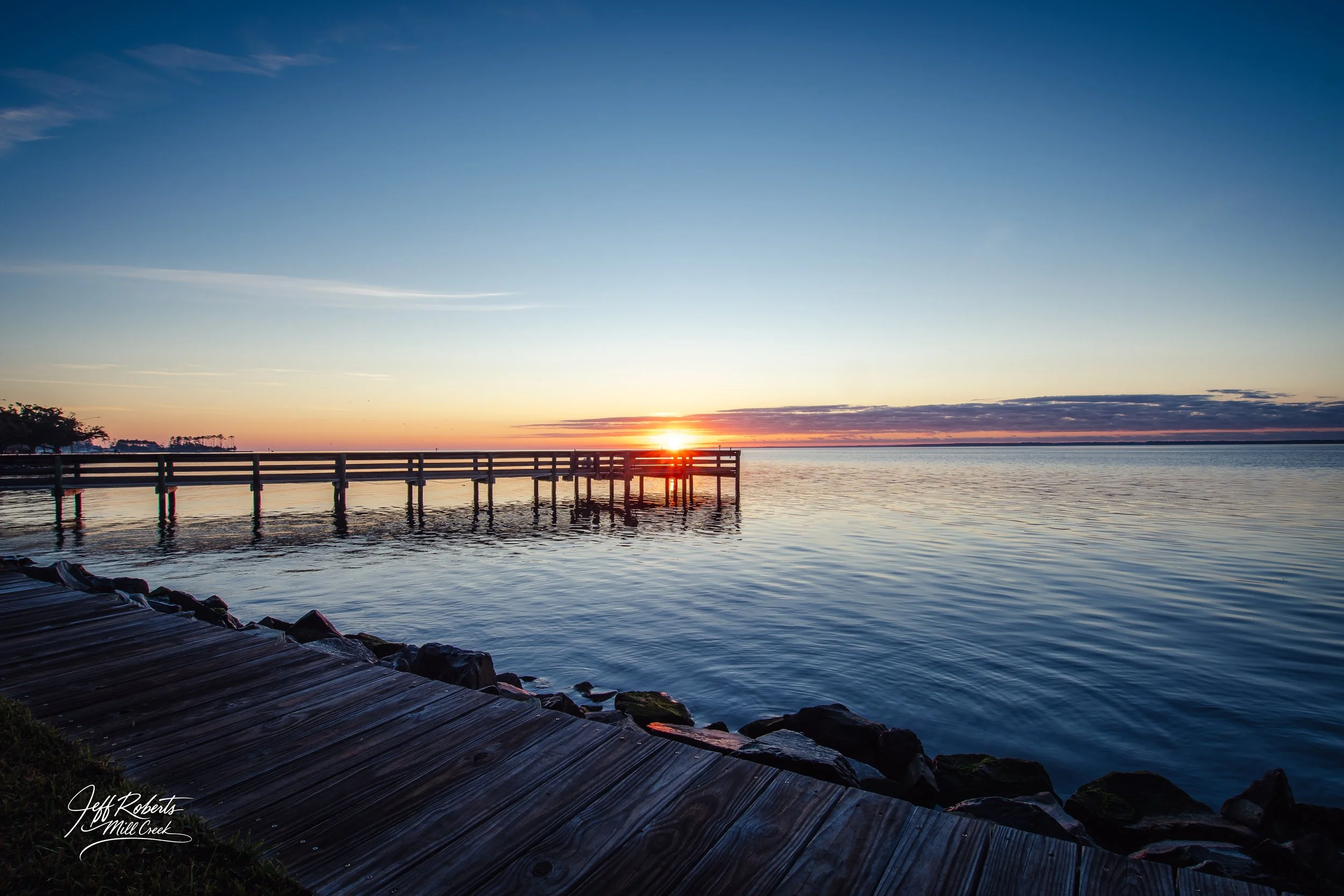 Sunset over a calm body of water with a wooden pier extending into the water and a wooden walkway along the shore.