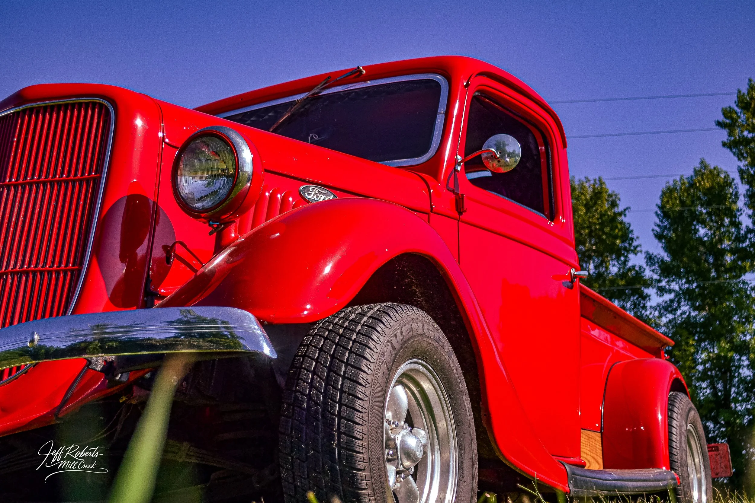 Close-up of a vintage red Ford truck parked outdoors with a clear blue sky and green trees in the background.