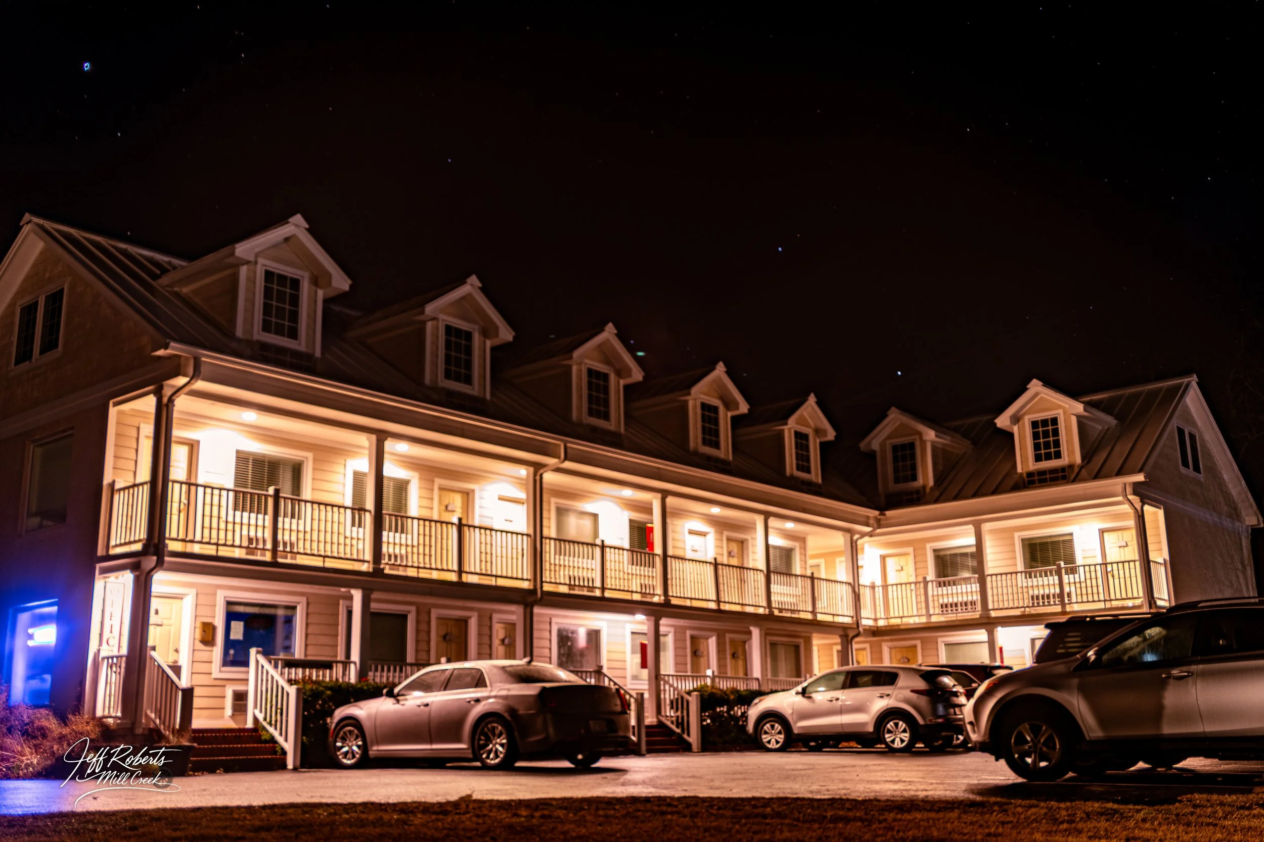 Nighttime view of a multi-story residential apartment building with exterior lighting, several parked cars in front, and a clear sky with visible stars.
