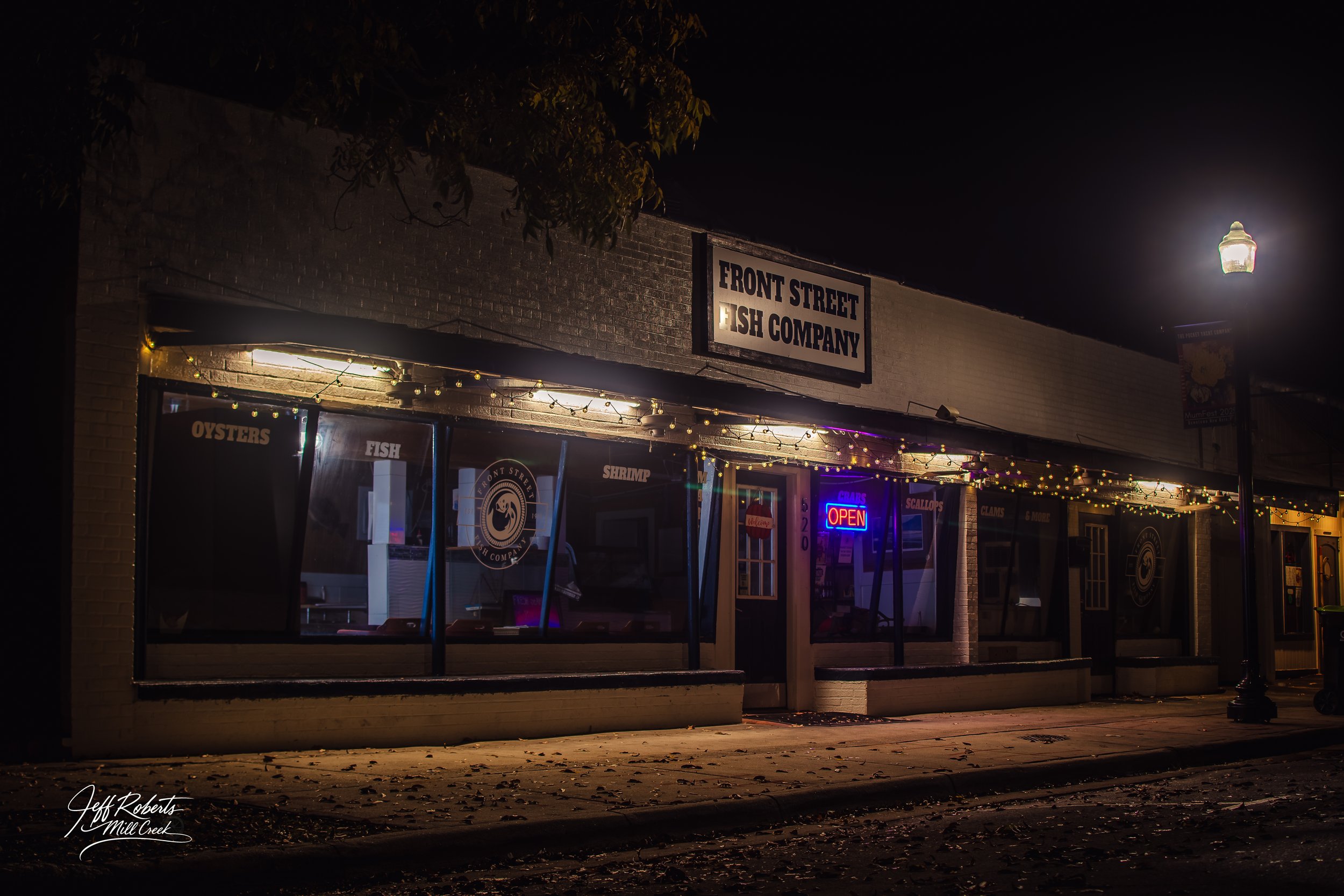 Night view of a seafood restaurant called Front Street Fish Company with a bright neon 'Open' sign in the window, illuminated outdoor string lights, and fallen leaves on the sidewalk.