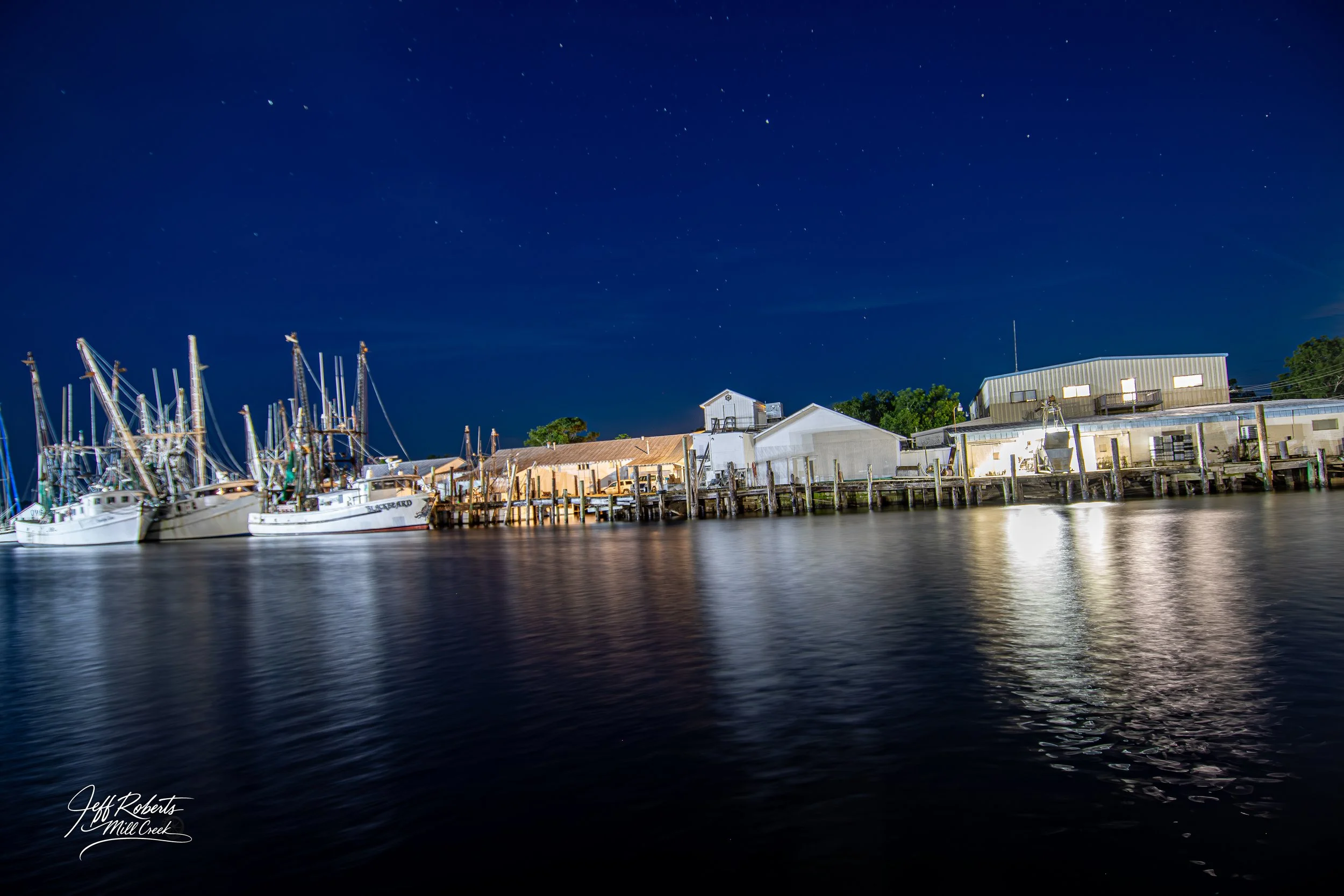 Nighttime view of a marina with several boats docked along wooden piers, reflected in the calm water, with a clear starry sky overhead and buildings in the background.