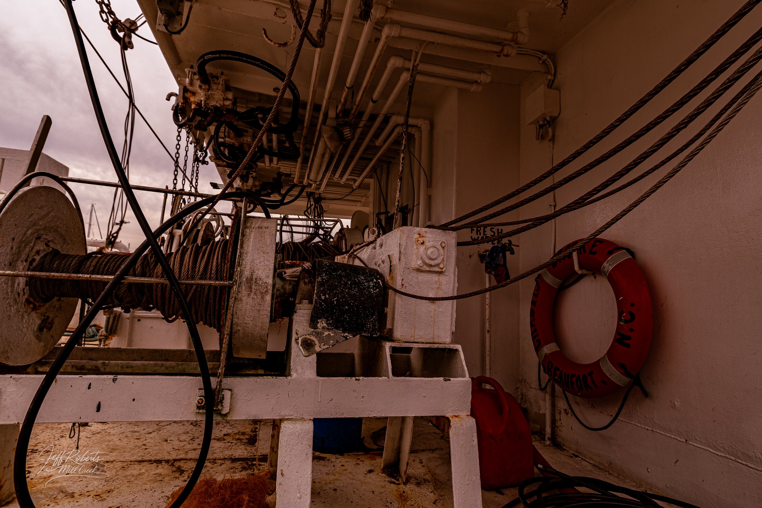 Close-up of a boat's equipment including a winch with wrapped rope, various pipes and hoses, a life buoy on the wall, and a fire extinguisher with a red container underneath, with a cloudy sky in the background.