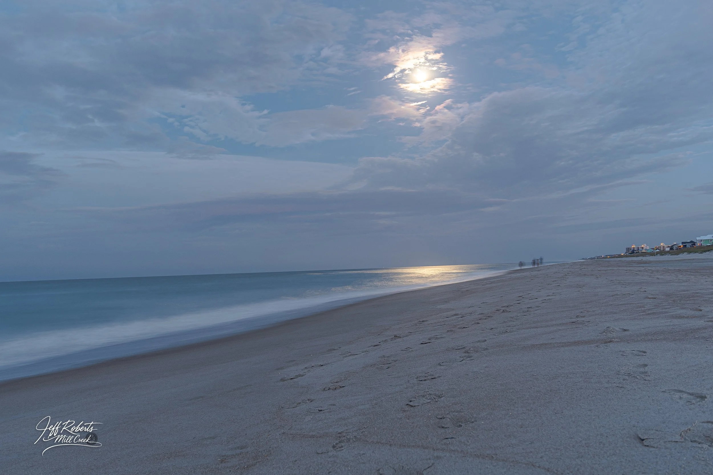 Calm beach with footprints in the sand, ocean waves, partly cloudy sky with sun peeking through clouds, and distant houses along the shoreline.