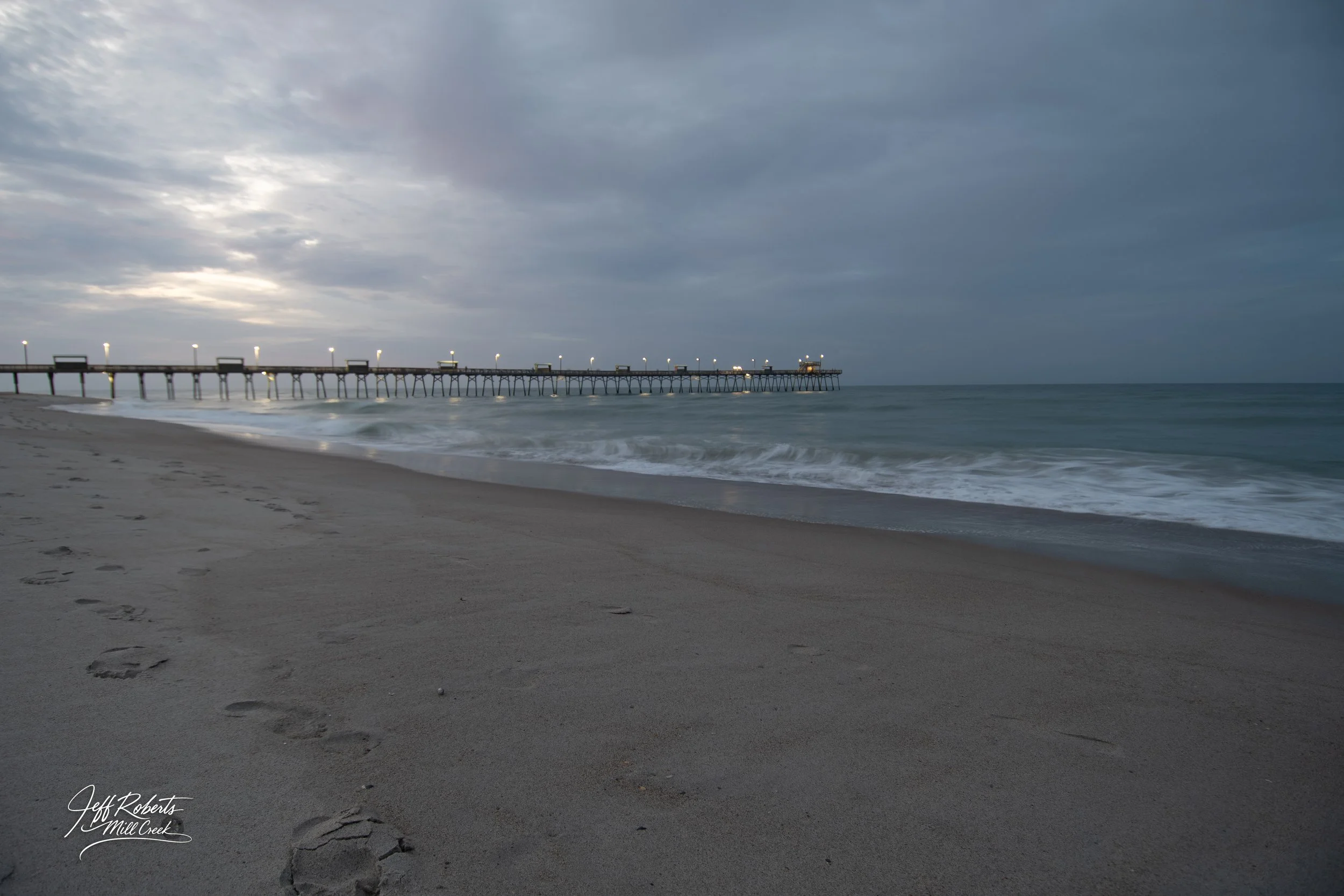 A beach with footprints in the sand, a pier extending into the ocean, and a cloudy sky overhead.