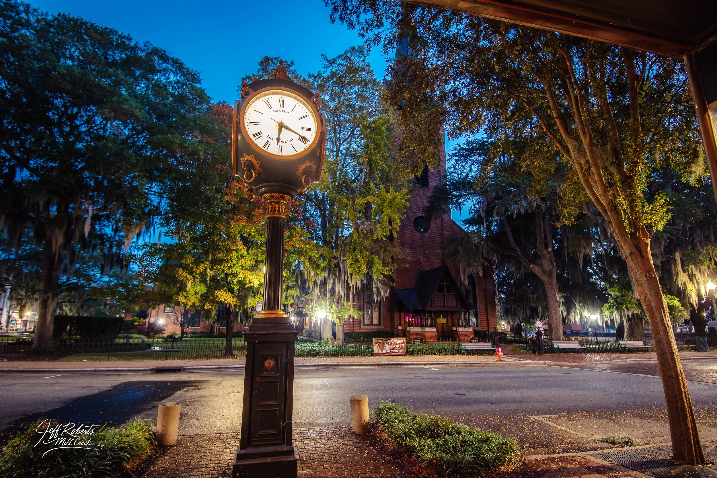 A vintage street clock showing approximately 5:20, located on a sidewalk in front of a historic brick church with large trees and hanging moss, illuminated during evening hours.