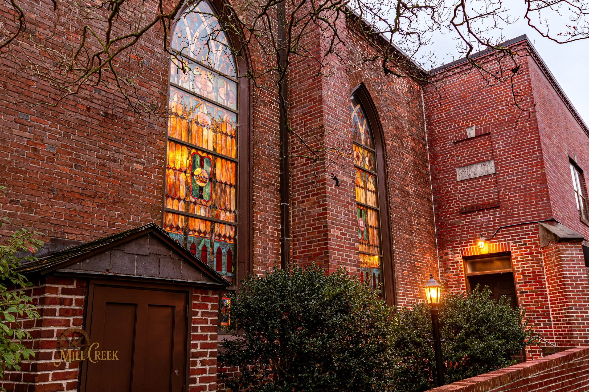 The exterior of a brick building with stained glass windows and two lit street lamps, with bushes and tree branches in the foreground.