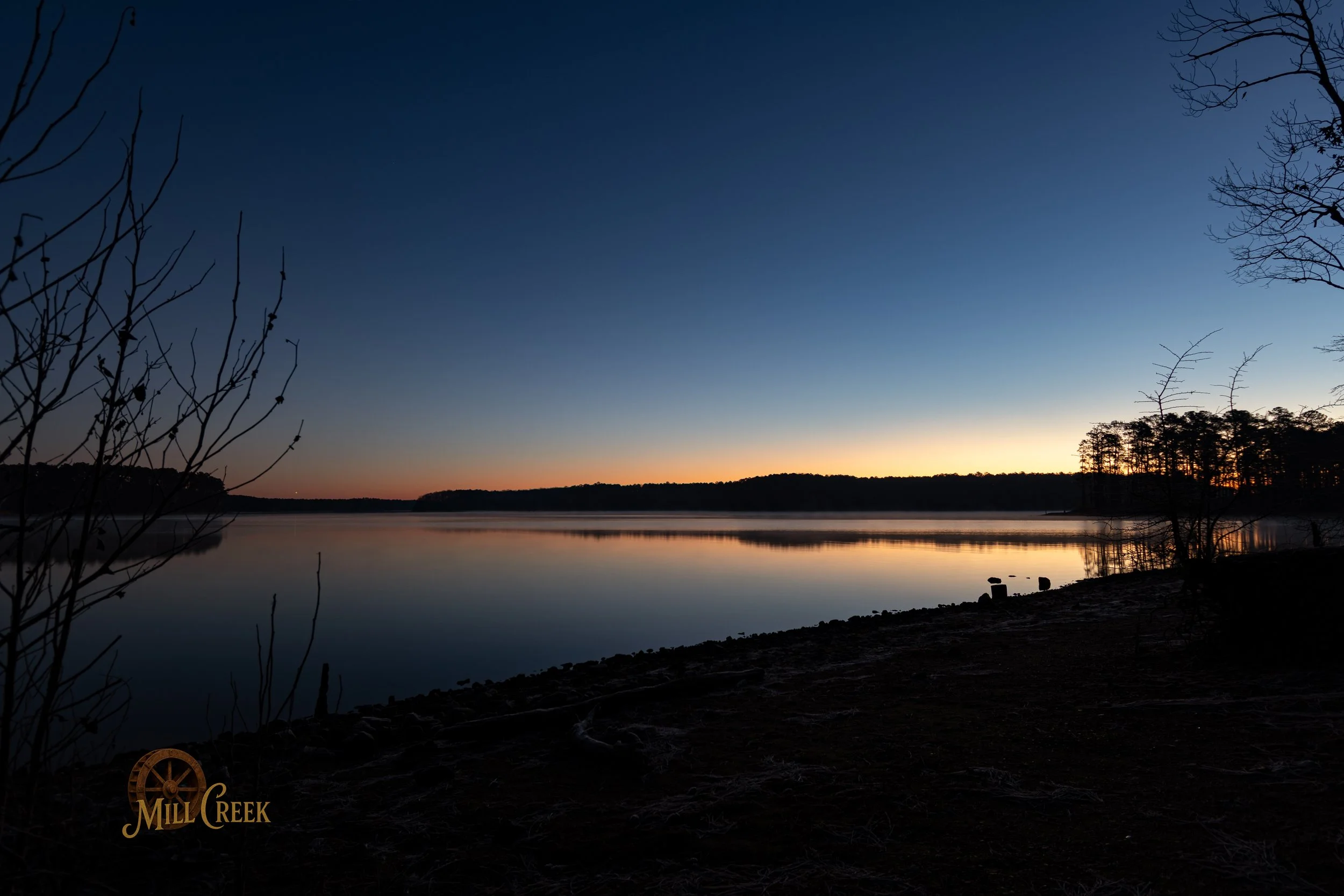 Serene lake at sunset with calm water reflecting the darkening sky, silhouetted trees along the shore, and a clear horizon with faint orange glow.