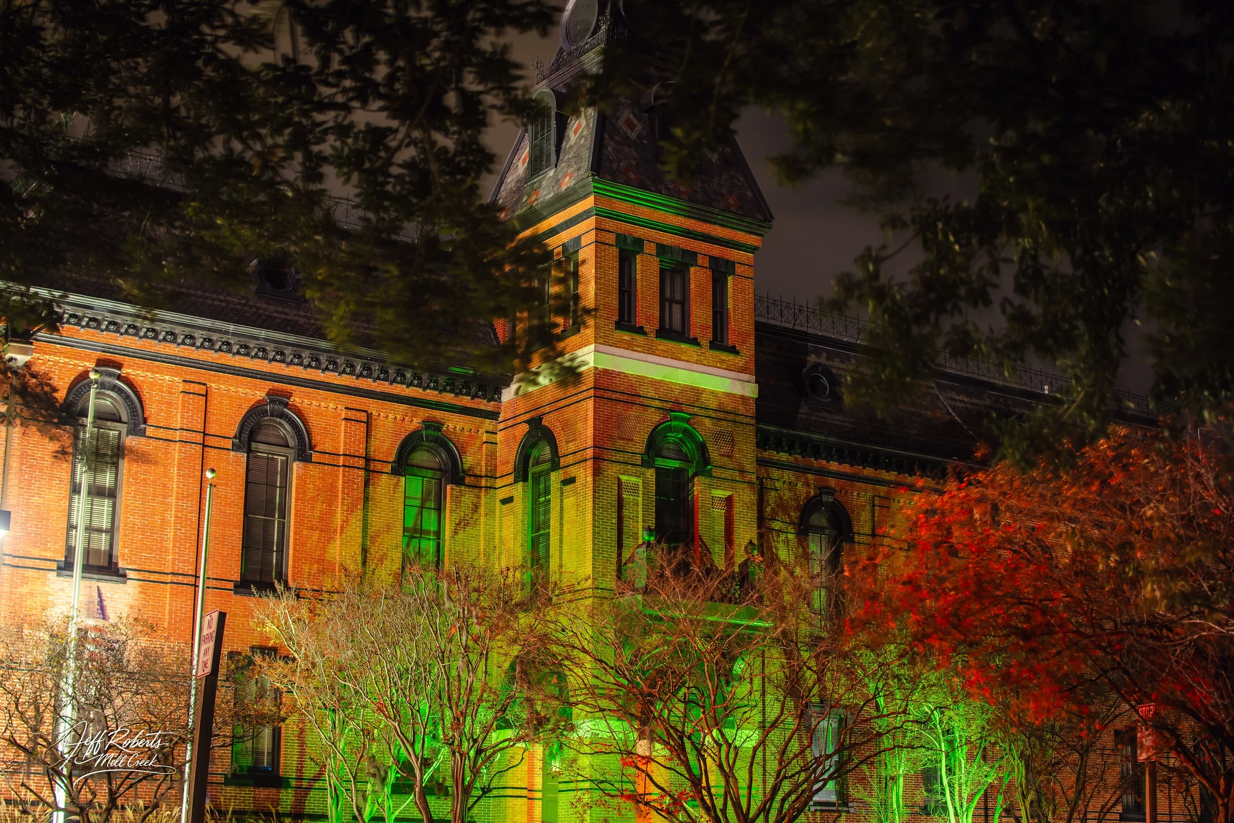 Historic brick building illuminated with green and red lights at night surrounded by trees with fall foliage.