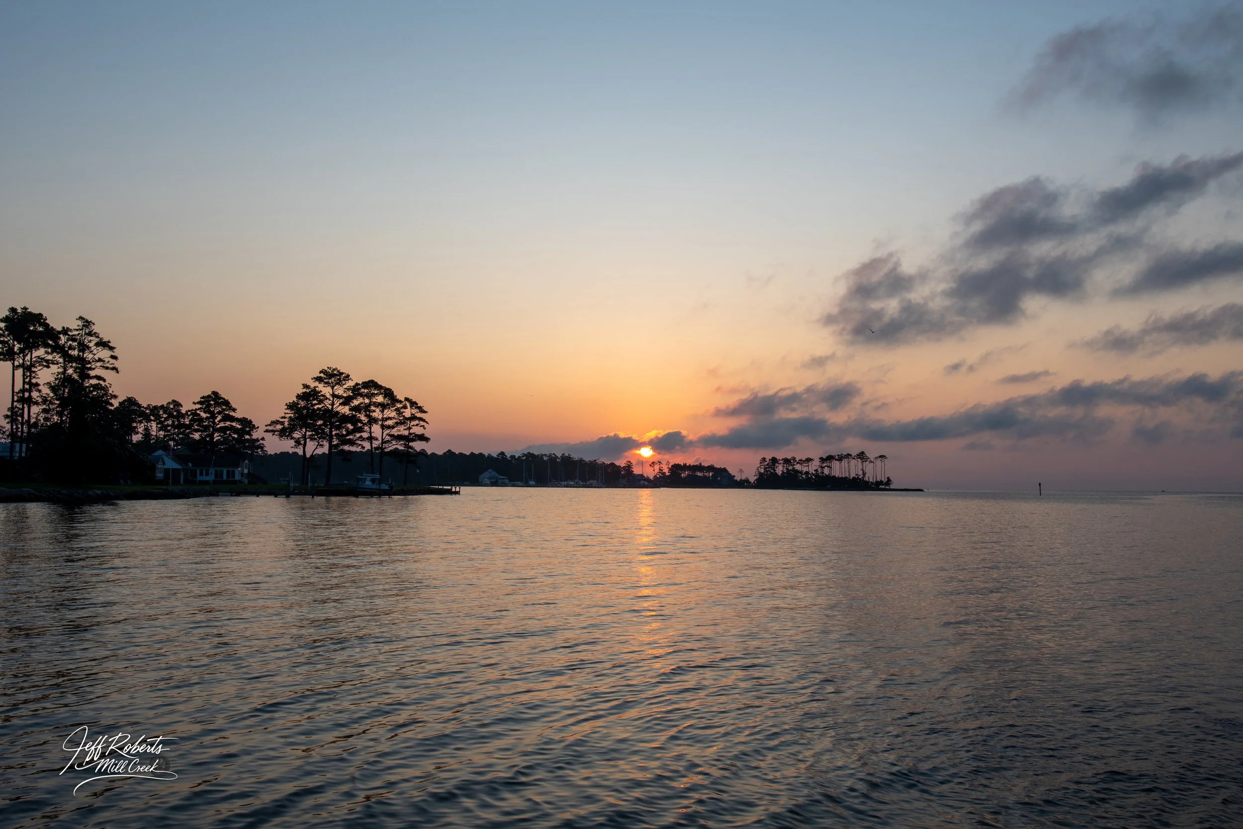 Sunset over a calm water body with trees and houses along the shoreline, partly cloudy sky.