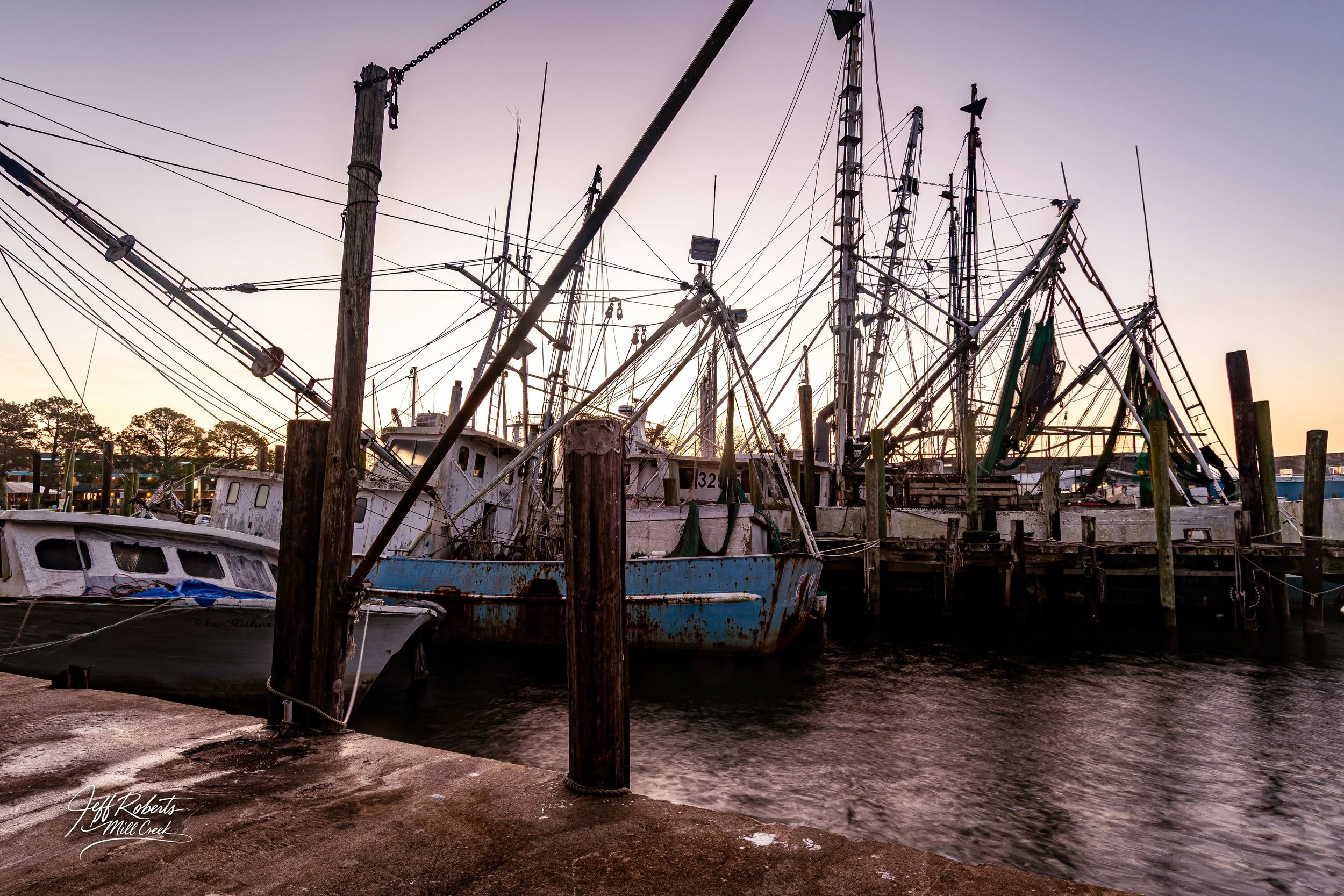 A collection of old, weathered boats docked at a pier during sunset, with a sky in shades of purple and pink and trees in the background.