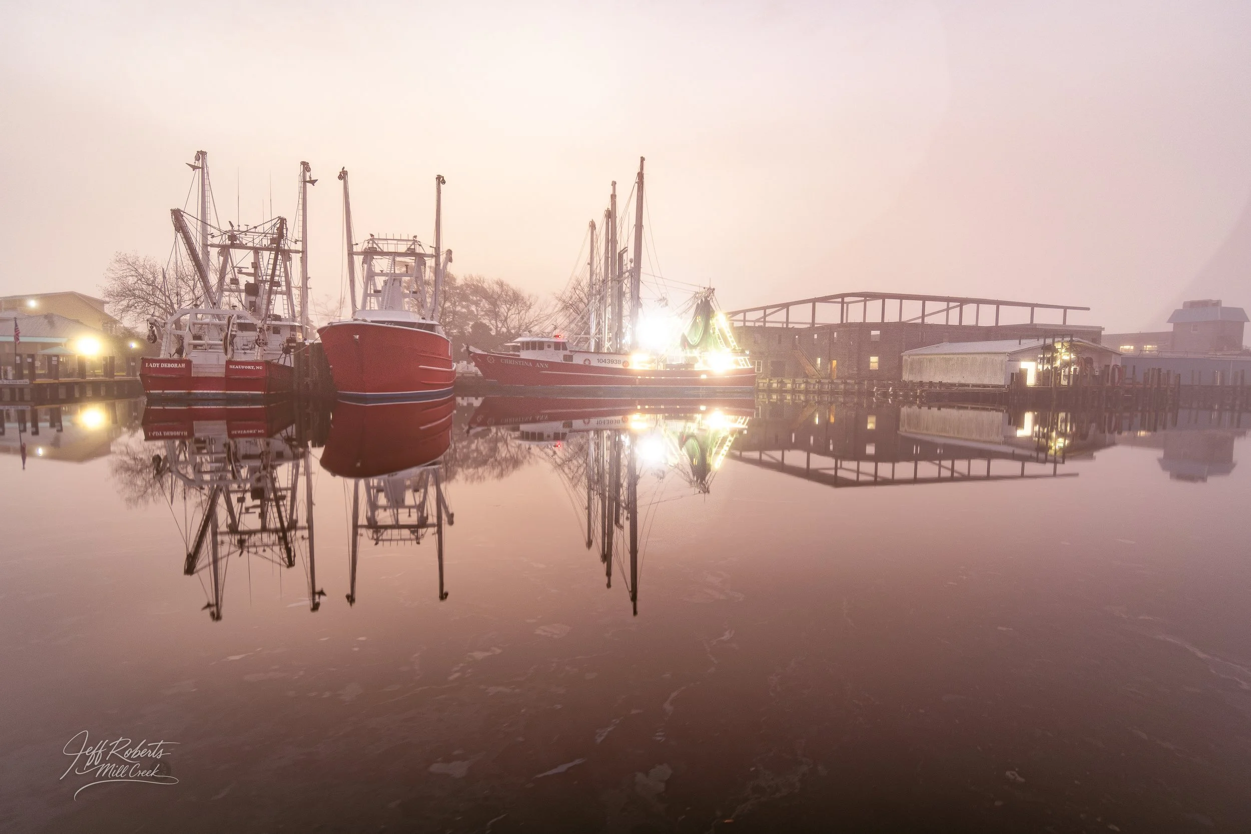 Boats docked at a marina with their reflections on the calm water, foggy background, and soft lighting.