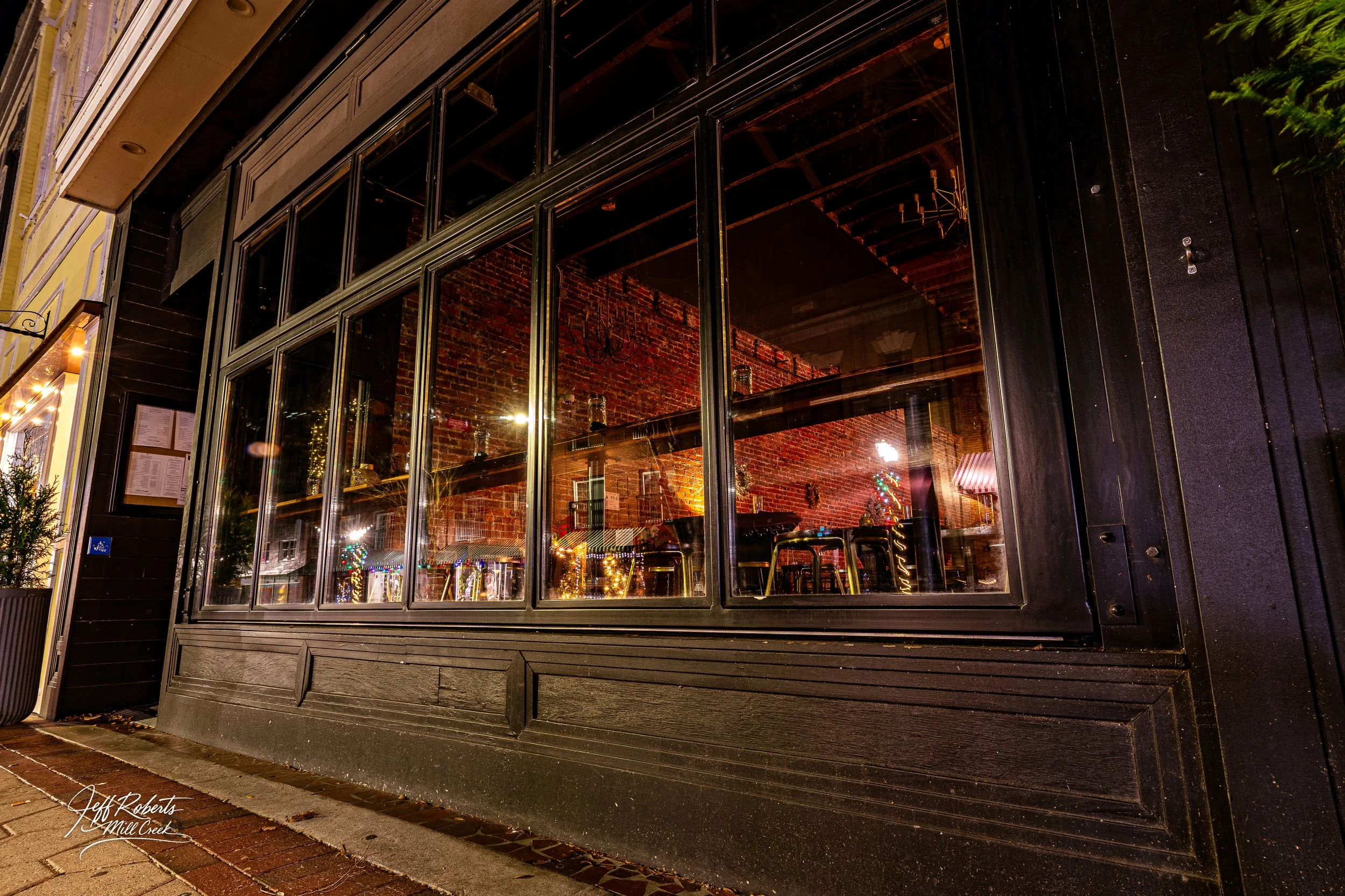 Night view of a restaurant or cafe through large glass windows showing a cozy, decorated interior with brick walls and tables.