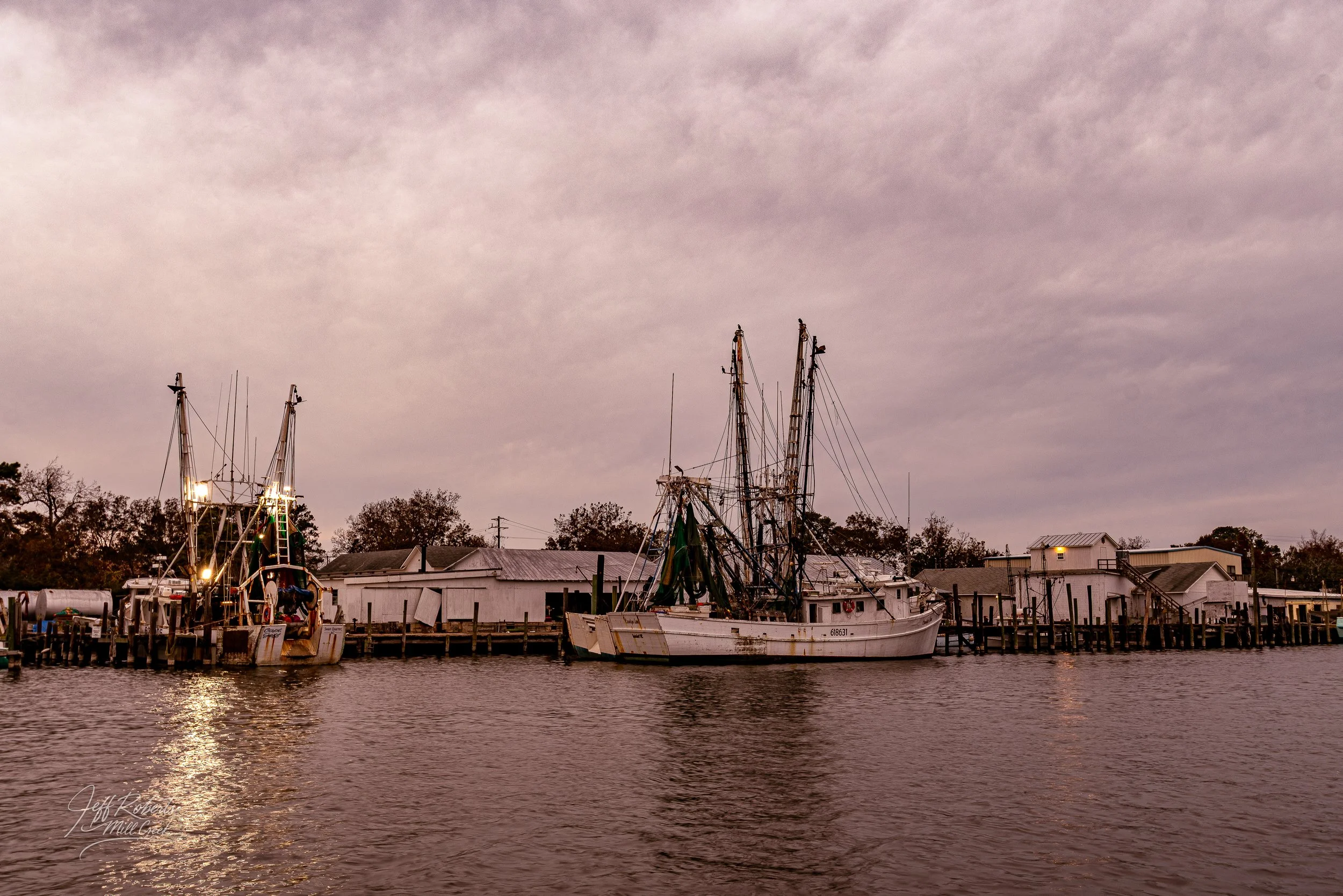 Fishing boats docked at a pier near a small harbor under a cloudy sky during sunset.