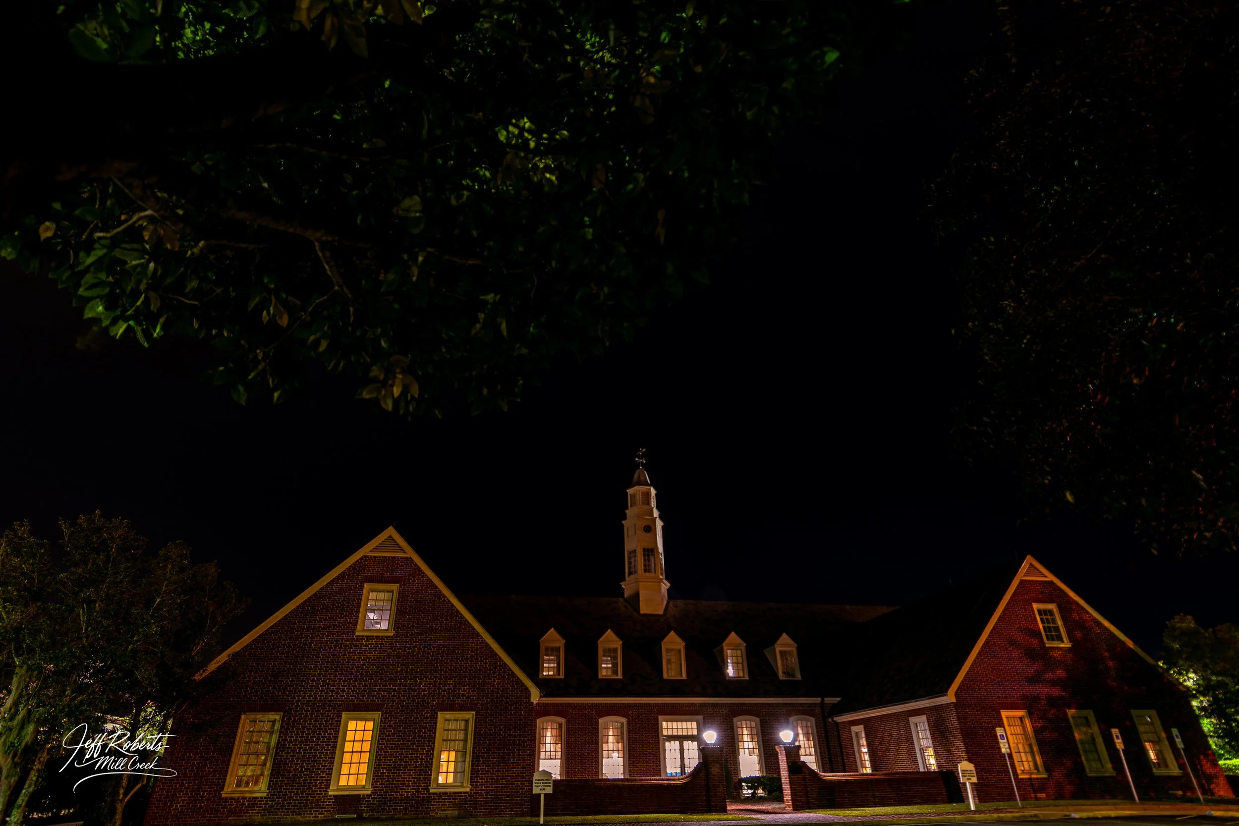 Nighttime view of a brick building with illuminated windows and a steeple, partially obscured by trees.