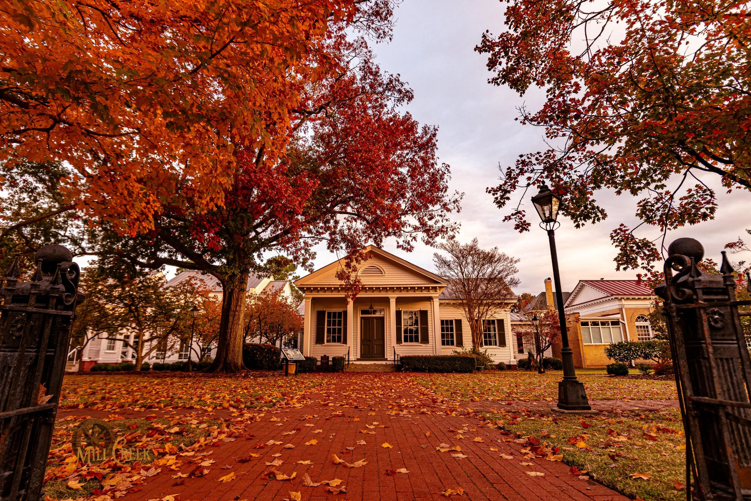 A historic white house with a dark door, surrounded by autumn trees with red and orange leaves, and a brick pathway with fallen leaves leading up to it, in a park-like setting.