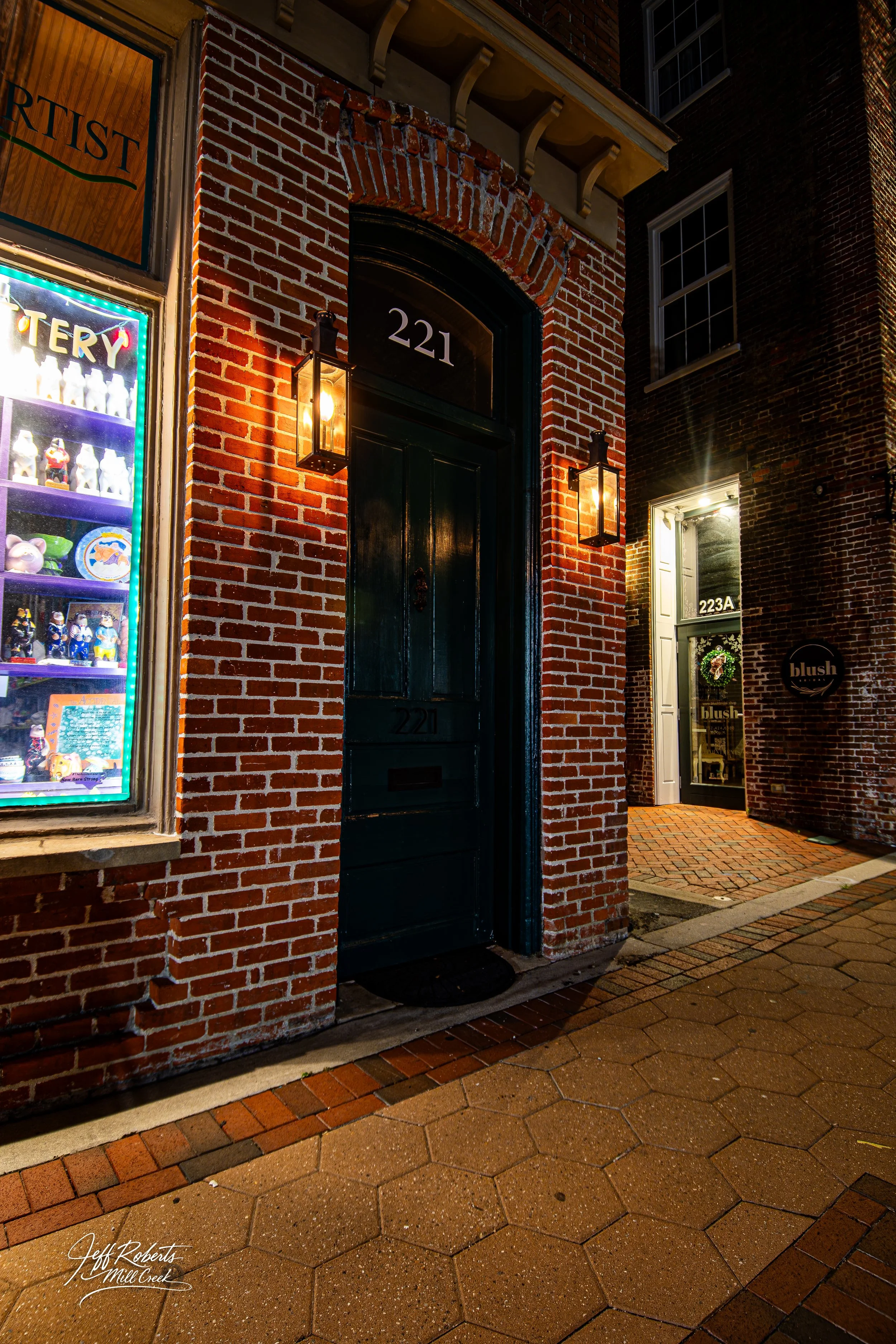 Nighttime view of a brick building with address 221, featuring a dark door with wall-mounted lanterns on either side, a window displaying figurines and collectibles on the left, and another door with a wreath on the right.