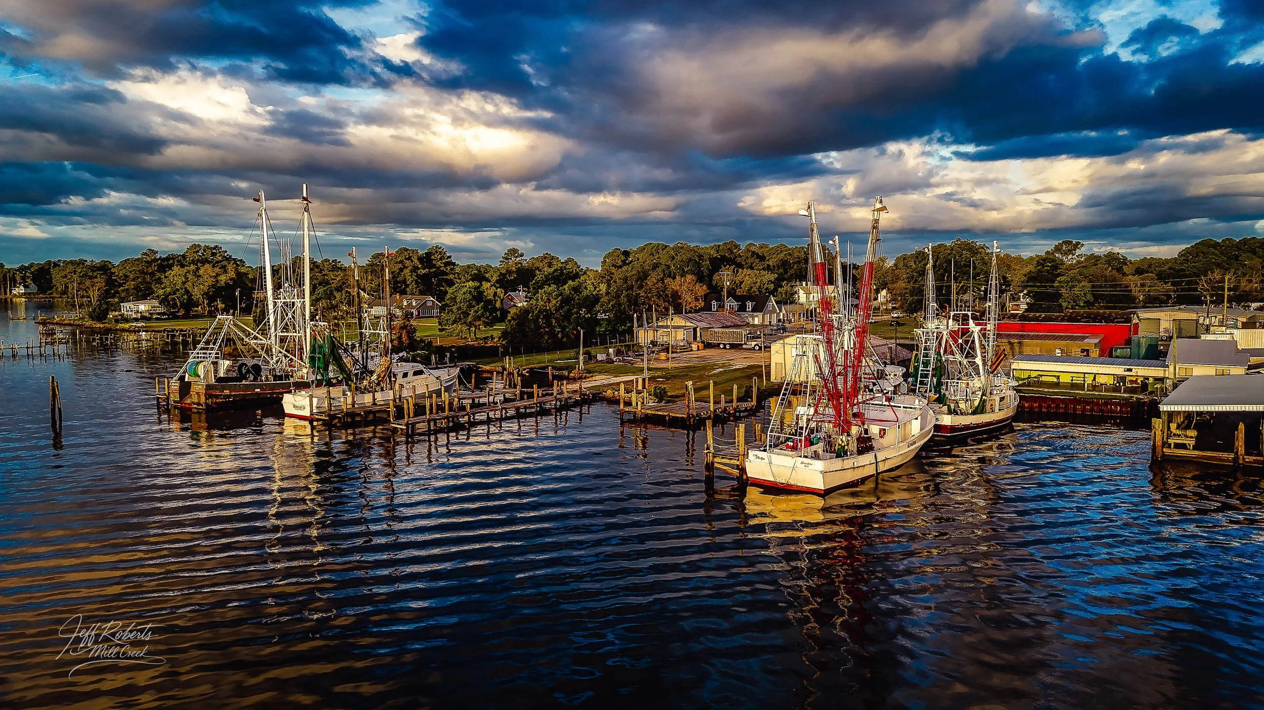 A marina with several boats docked at the pier during sunset, with clouds in the sky and trees in the background.