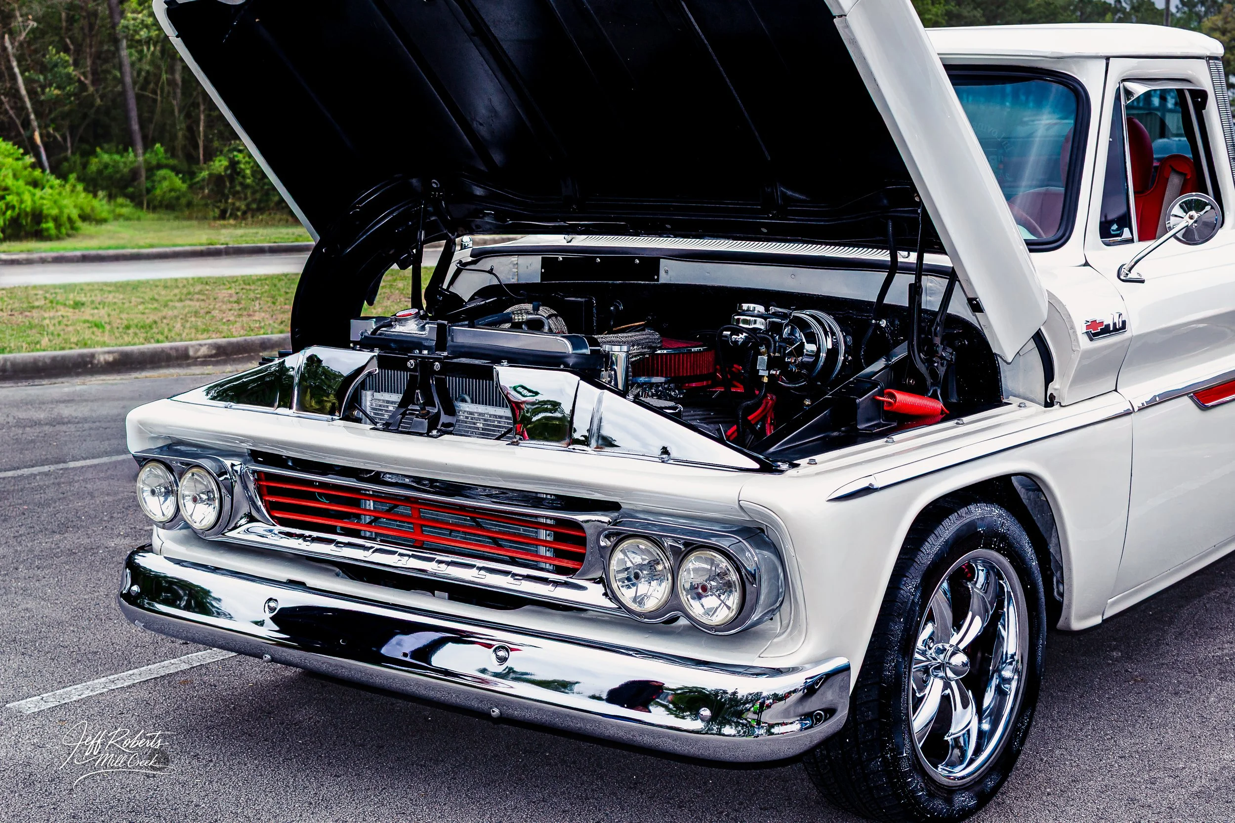 A vintage white pickup truck with its hood open, revealing a custom engine with chrome and red components, parked on pavement with a grassy area and trees in the background.
