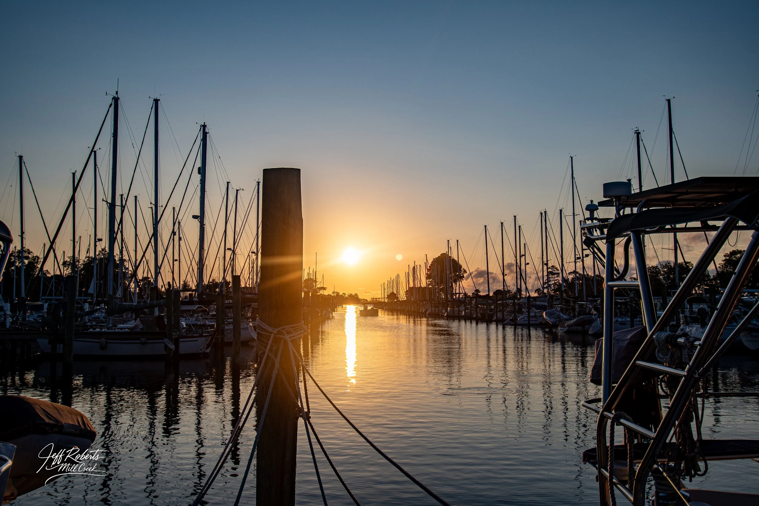 Sunset over a marina with sailboats and yachts docked along the pier, reflection of the sunset on the water, and a few trees in the background.