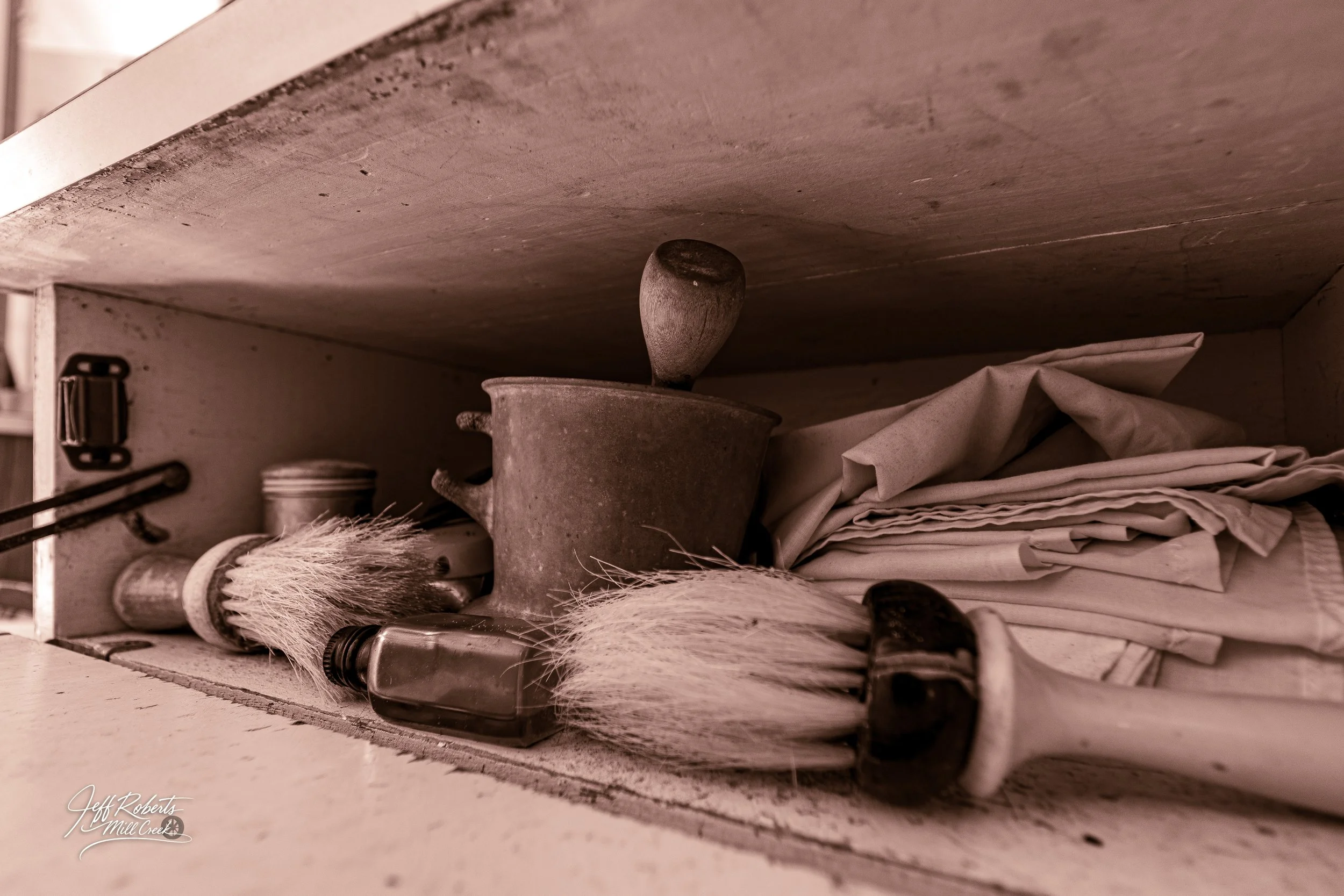Open kitchen cabinet shelf with vintage kitchen items including a metal handheld mixer, a small glass bottle, a bristle brush, and a stack of folded cloths or linens.