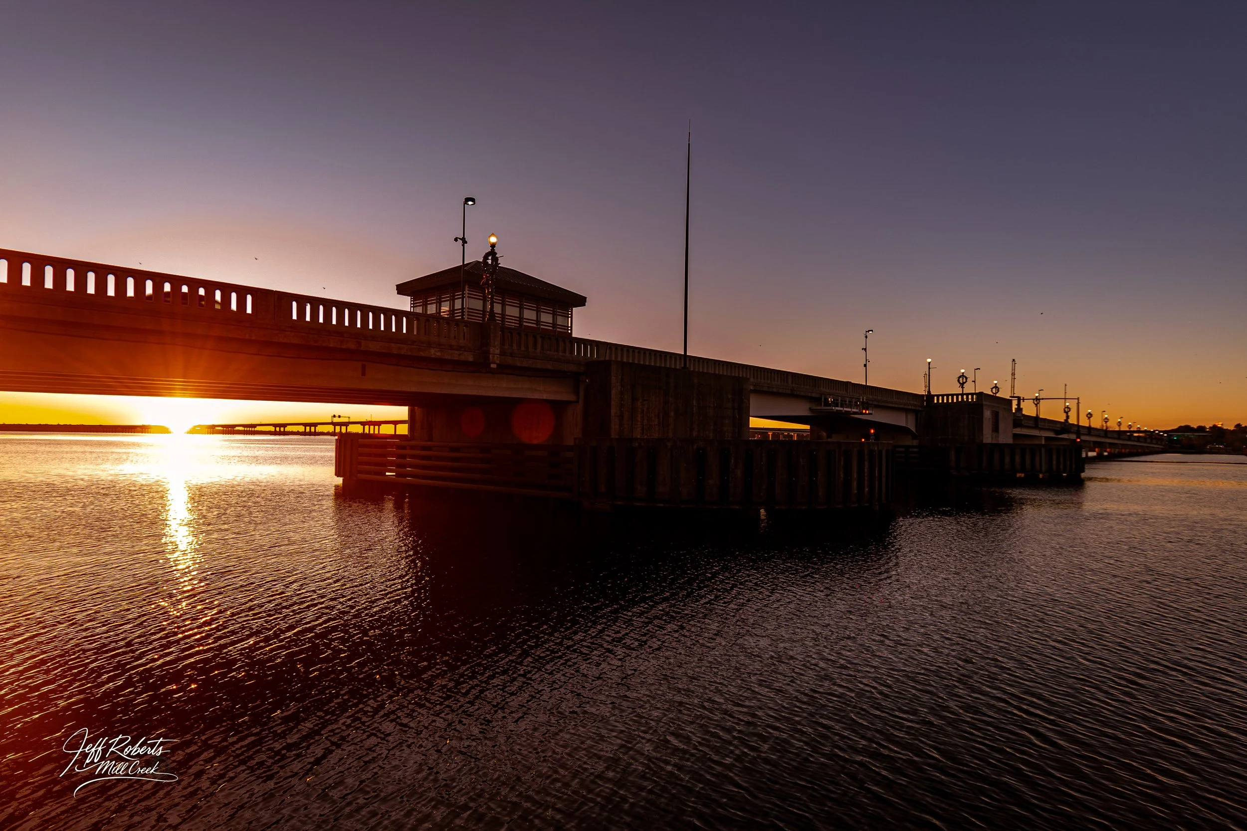 Sunset view of a wooden pier extending into water, with street lamps and a small building on the pier, reflected in the water.