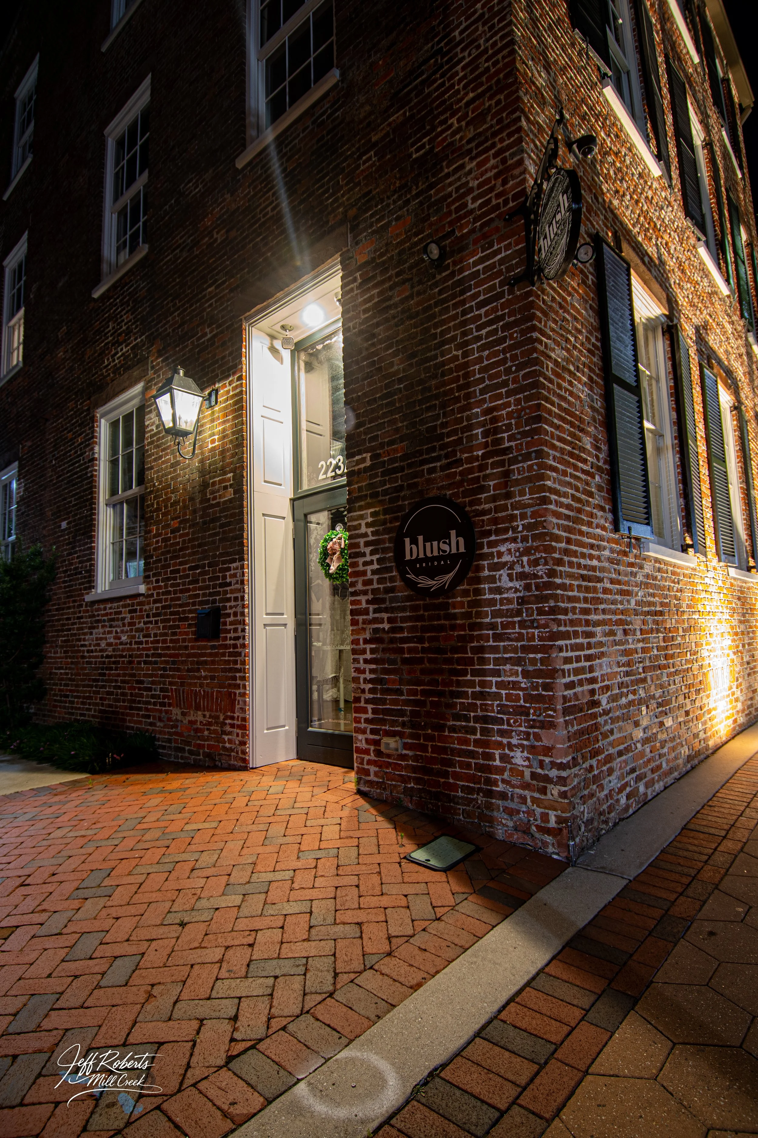 Nighttime view of a brick building with an illuminated entrance and a sign that reads 'blush' for a boutique. Brick walkway leading to the entrance, with streetlights casting warm light.