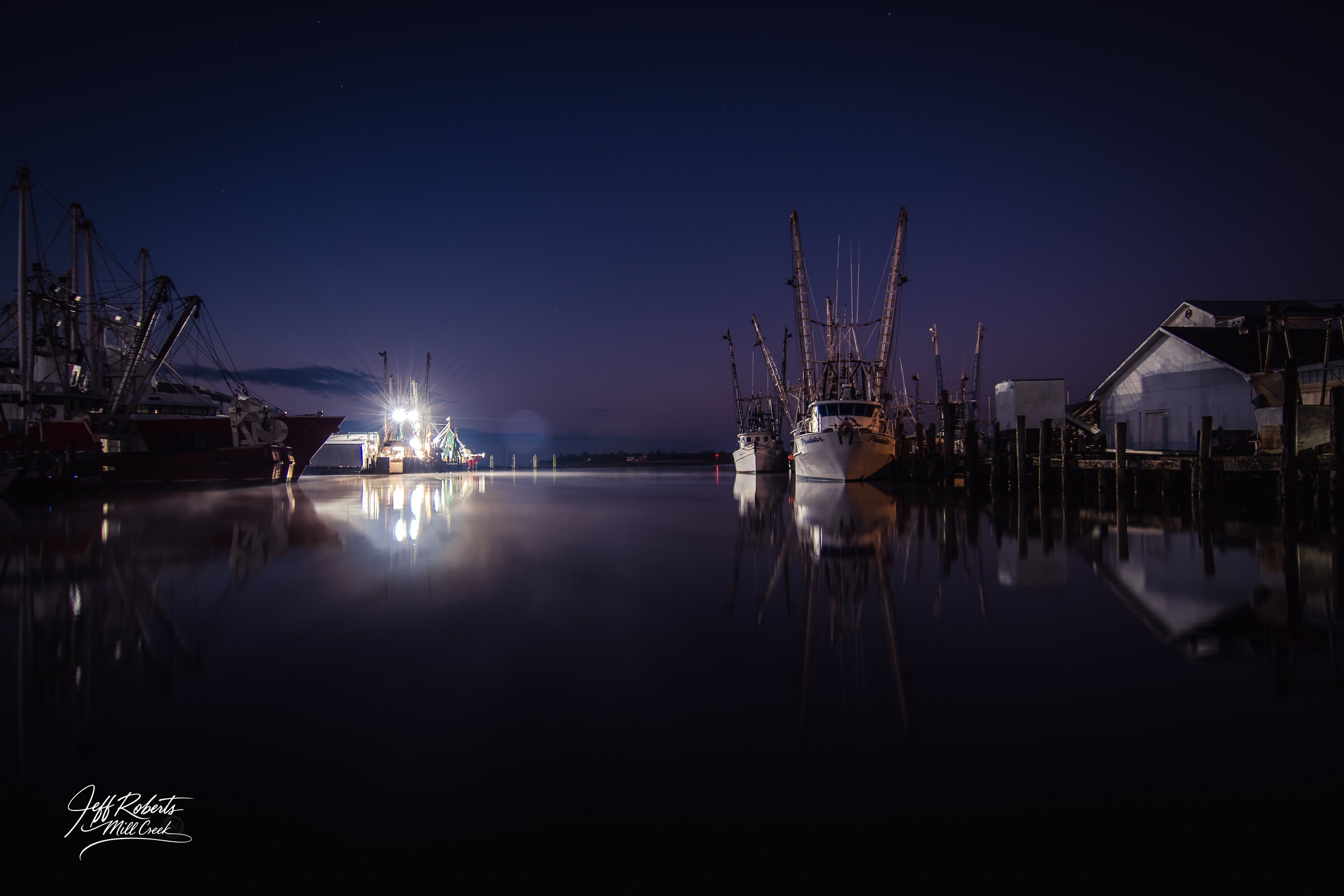 Nighttime view of a harbor with boats docked along the pier, reflected in the calm water, with a dark sky and a few stars visible.