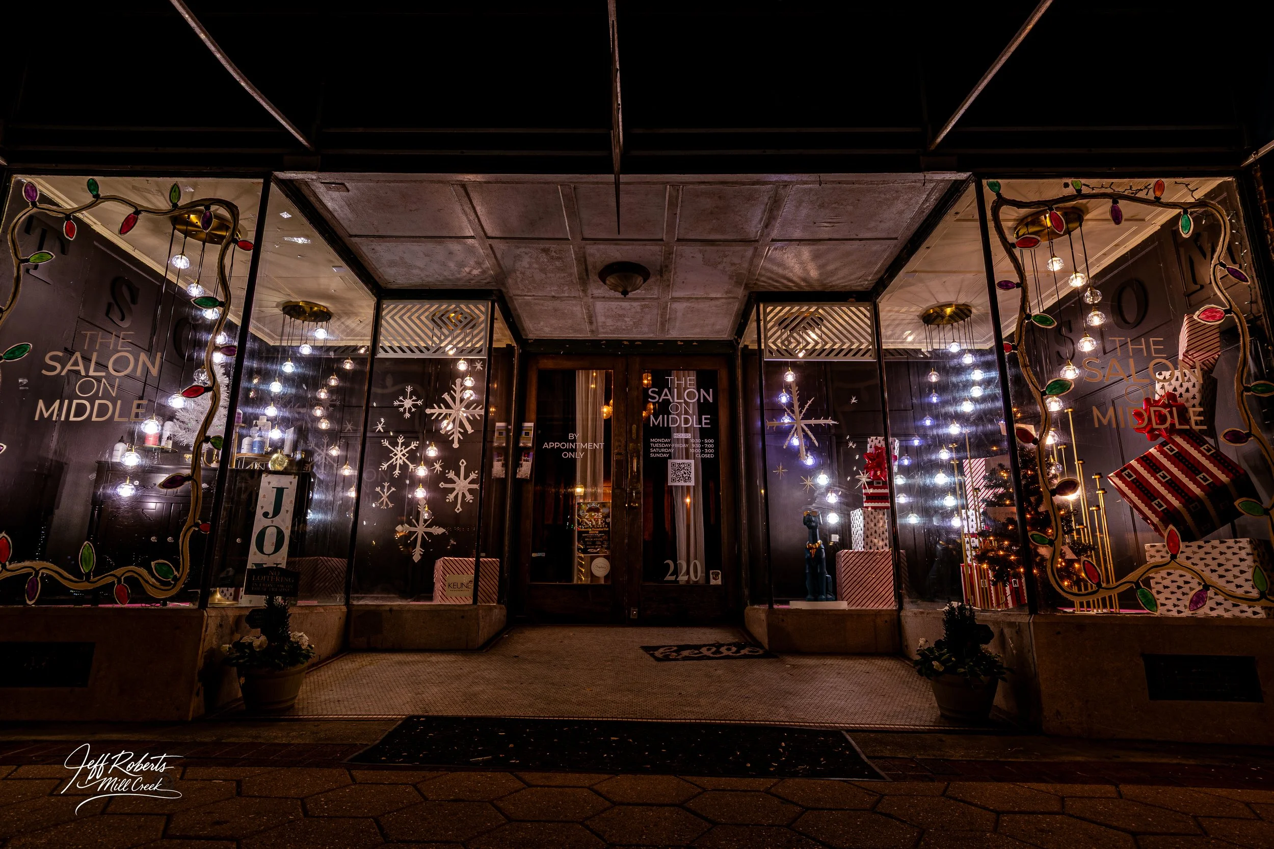 Storefront decorated for Christmas with holiday lights, snowflake window decals, wrapped presents, and a small Christmas tree inside