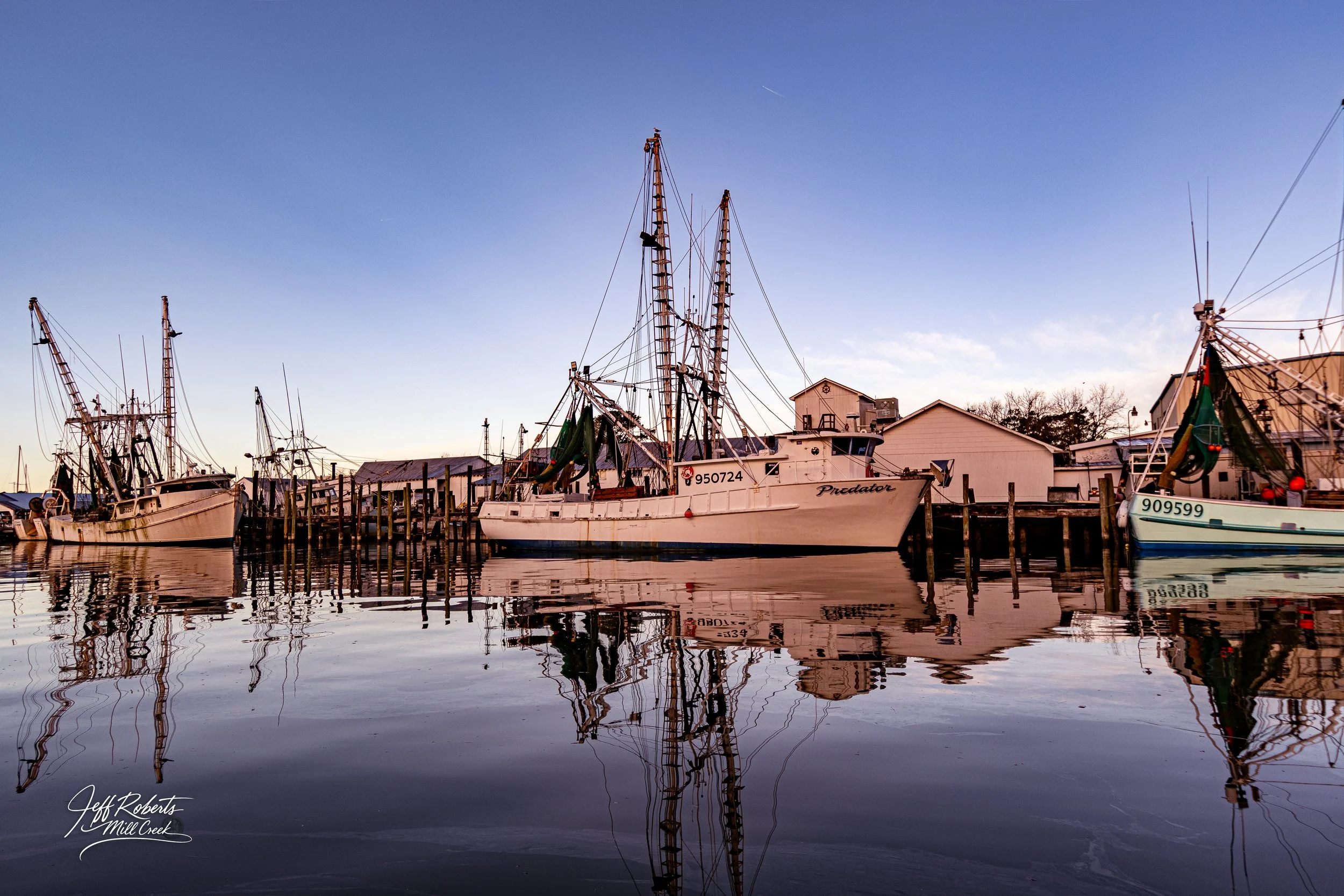 Boats docked at a marina during sunset, with calm water reflecting the ships and a clear blue sky.