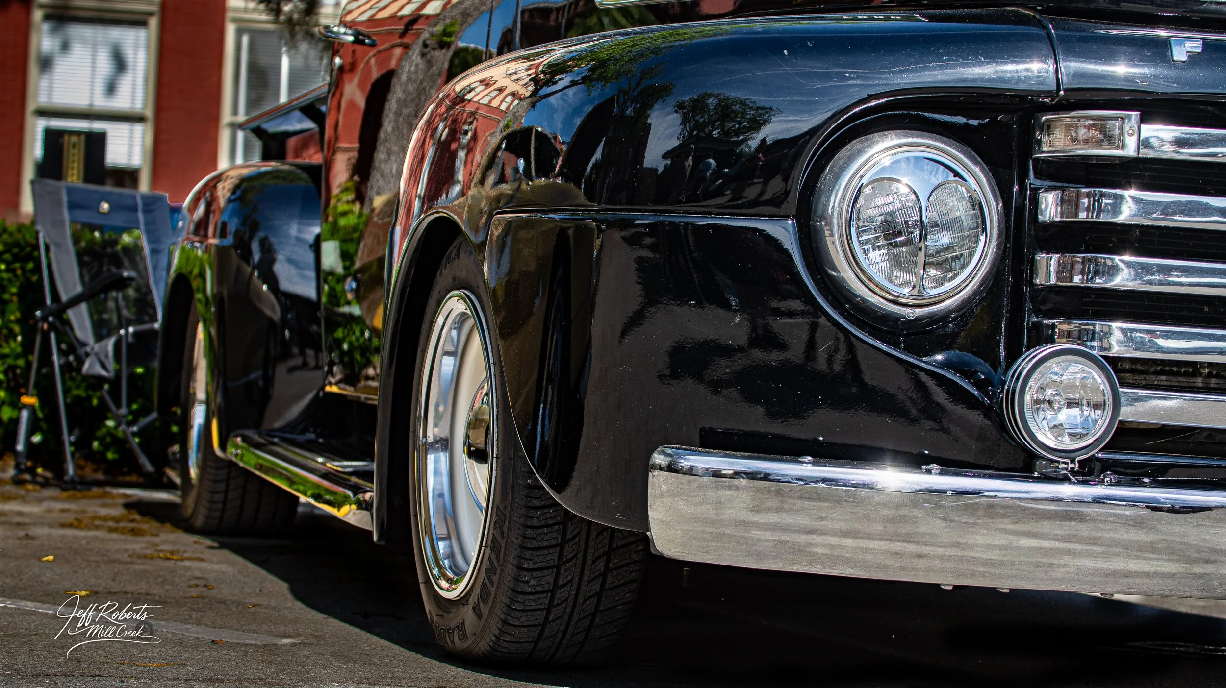 Close-up of a vintage black car parked on a street with a folding chair and green bushes in the background.