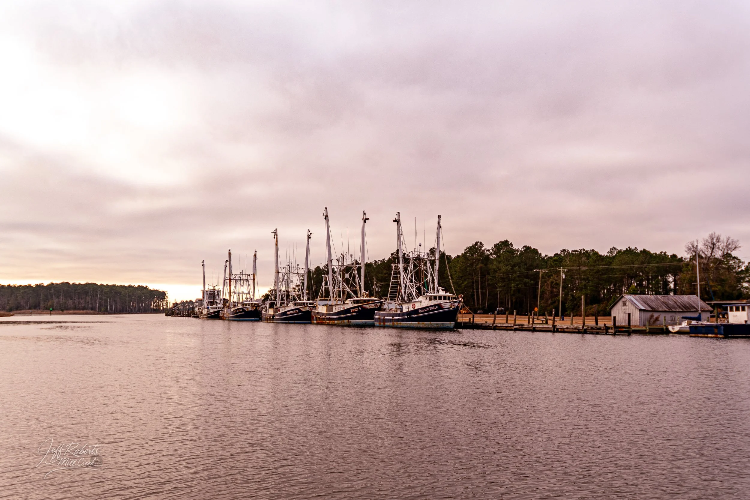 Marina with several docked boats on calm water, overcast sky, trees and small building in background.