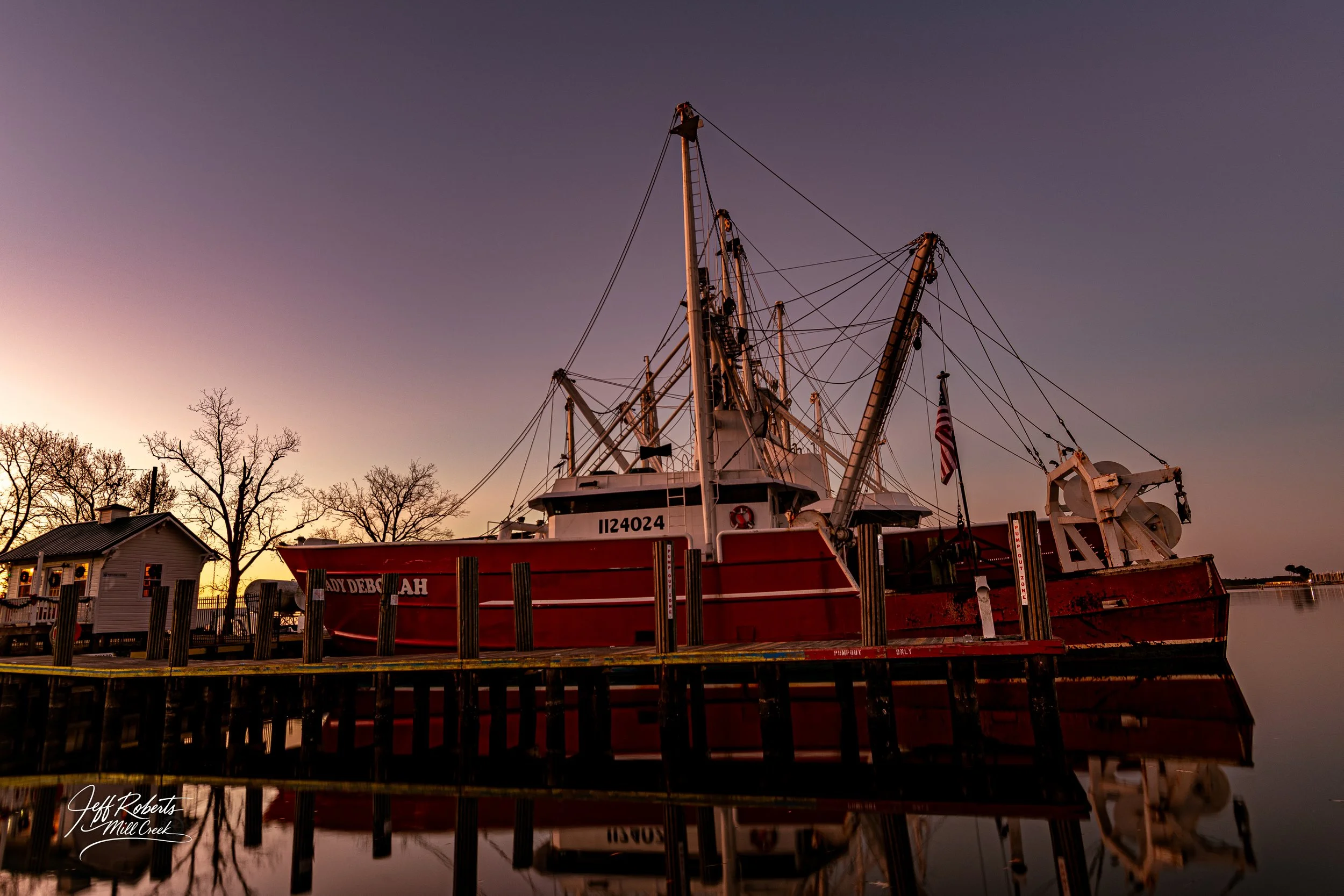 A red fireboat named LADY DEBBIE is docked at a waterfront, with reflections in the calm water during sunset or sunrise, silhouetted trees, and a small building nearby.