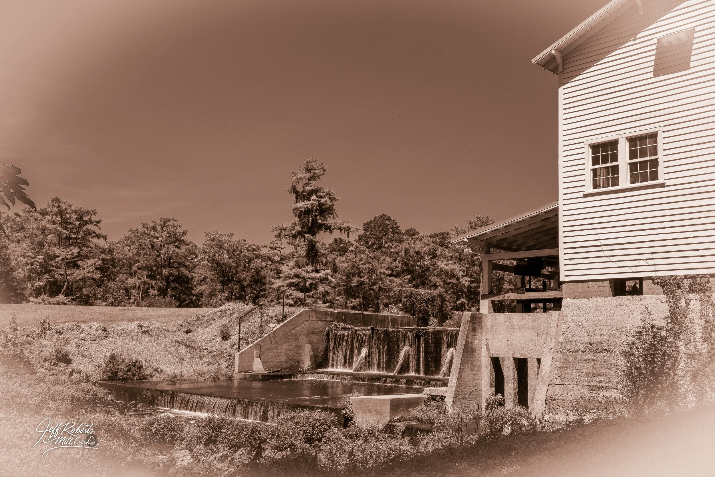 Old building with white siding and window, next to small dam with water cascading over ledges, surrounded by trees, sepia tone.