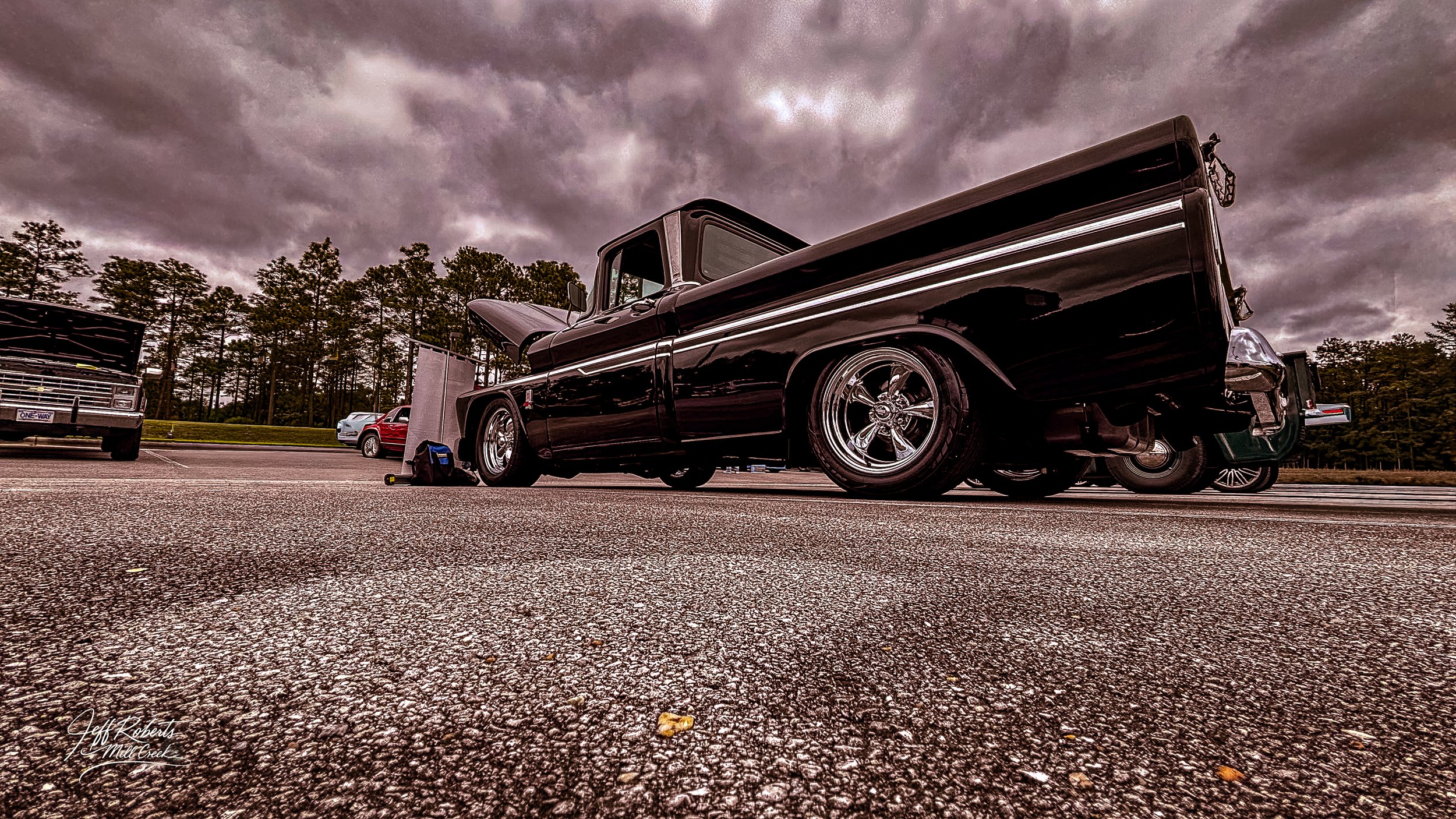 A black classic pickup truck with chrome wheels parked in a parking lot under a cloudy sky, with other vehicles and trees in the background.