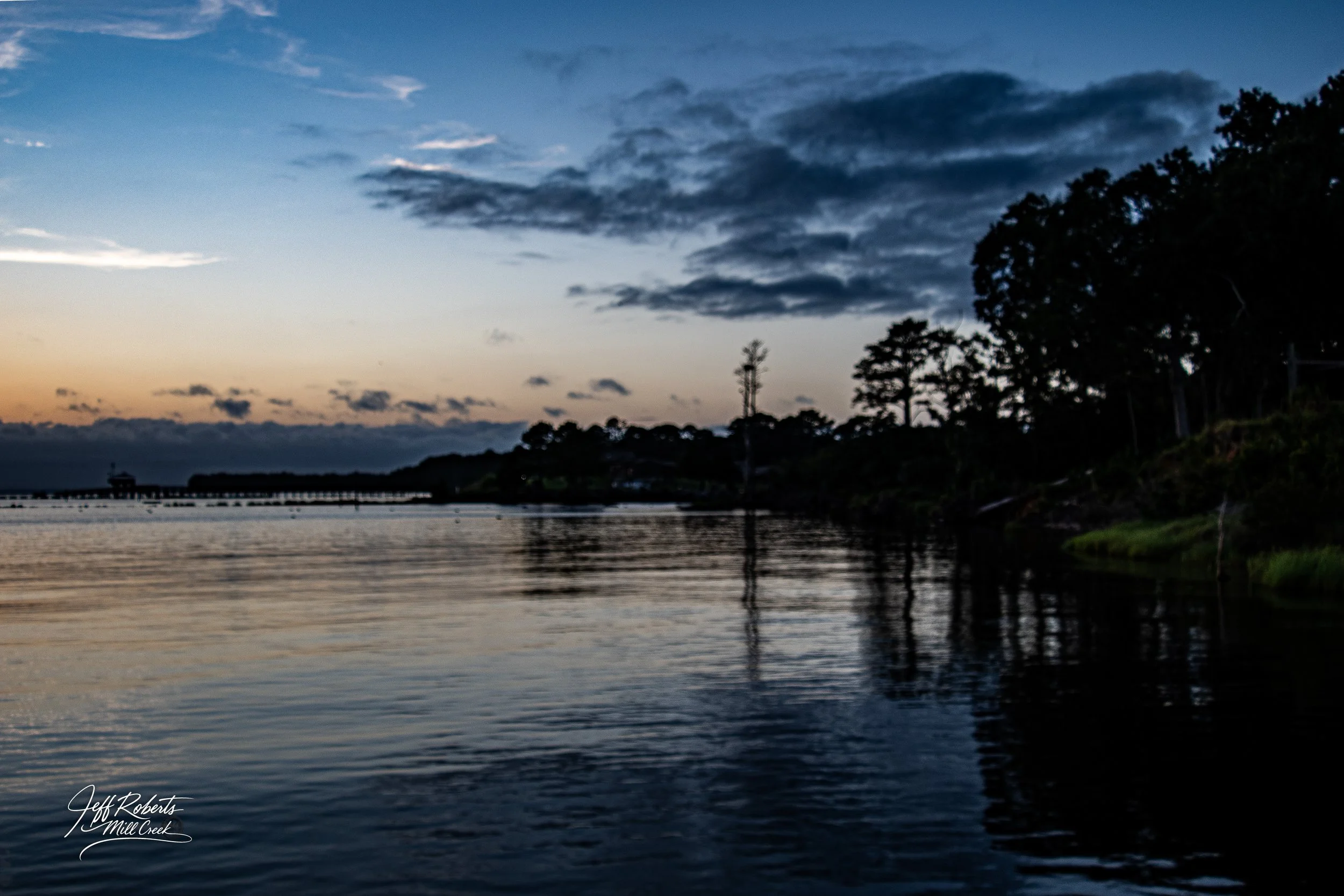A serene river scene at sunset with a wooden pier and trees along the shoreline, reflecting on the water under a partly cloudy sky.