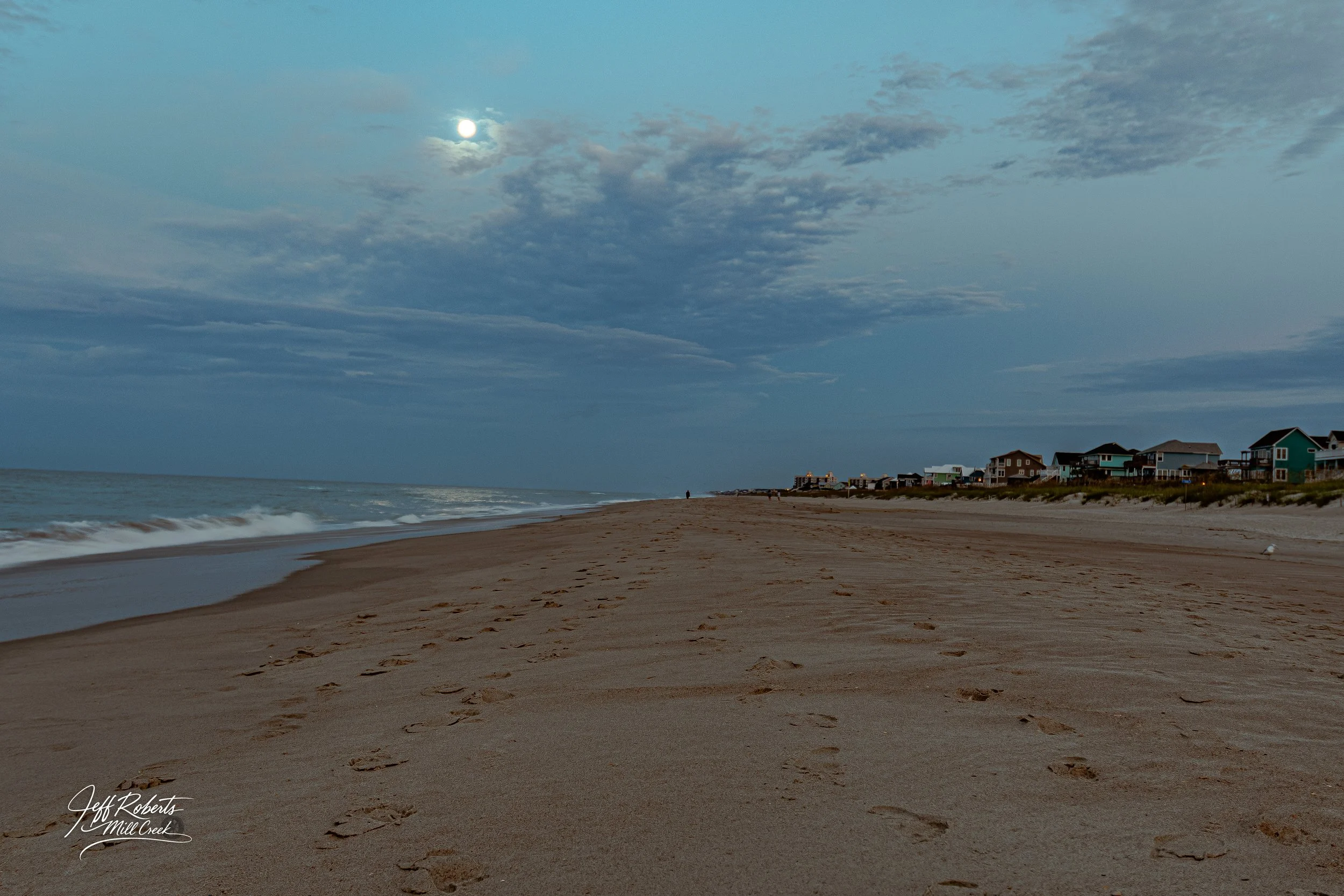 Beach at dusk with footprints in the sand, houses in the distance, a cloudy sky, and the moon visible.