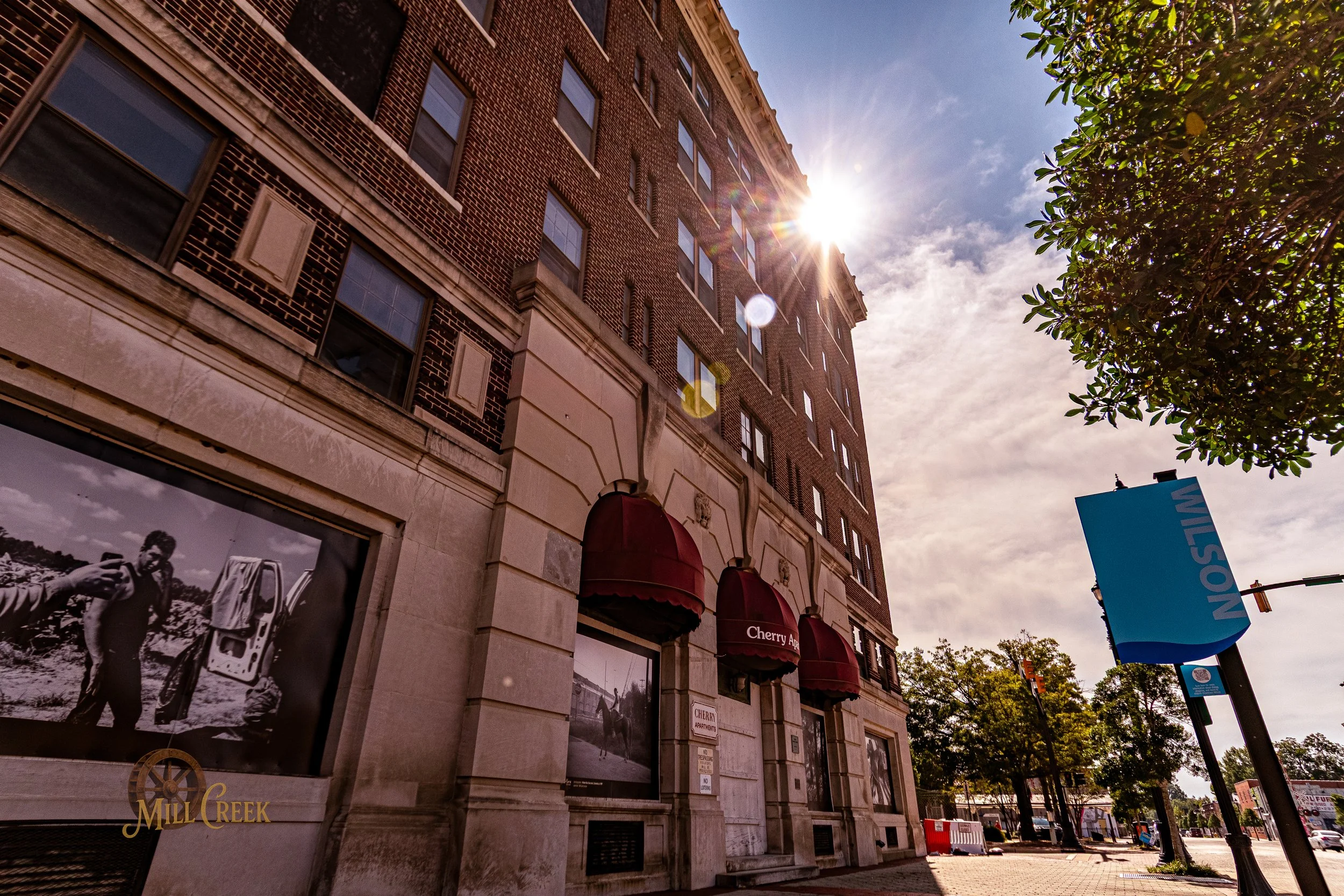A tall brick building with arched windows and red awnings on the ground floor. The sun is shining brightly from behind the building, creating a lens flare. There are trees and a street with parked cars and a sidewalk in the foreground.