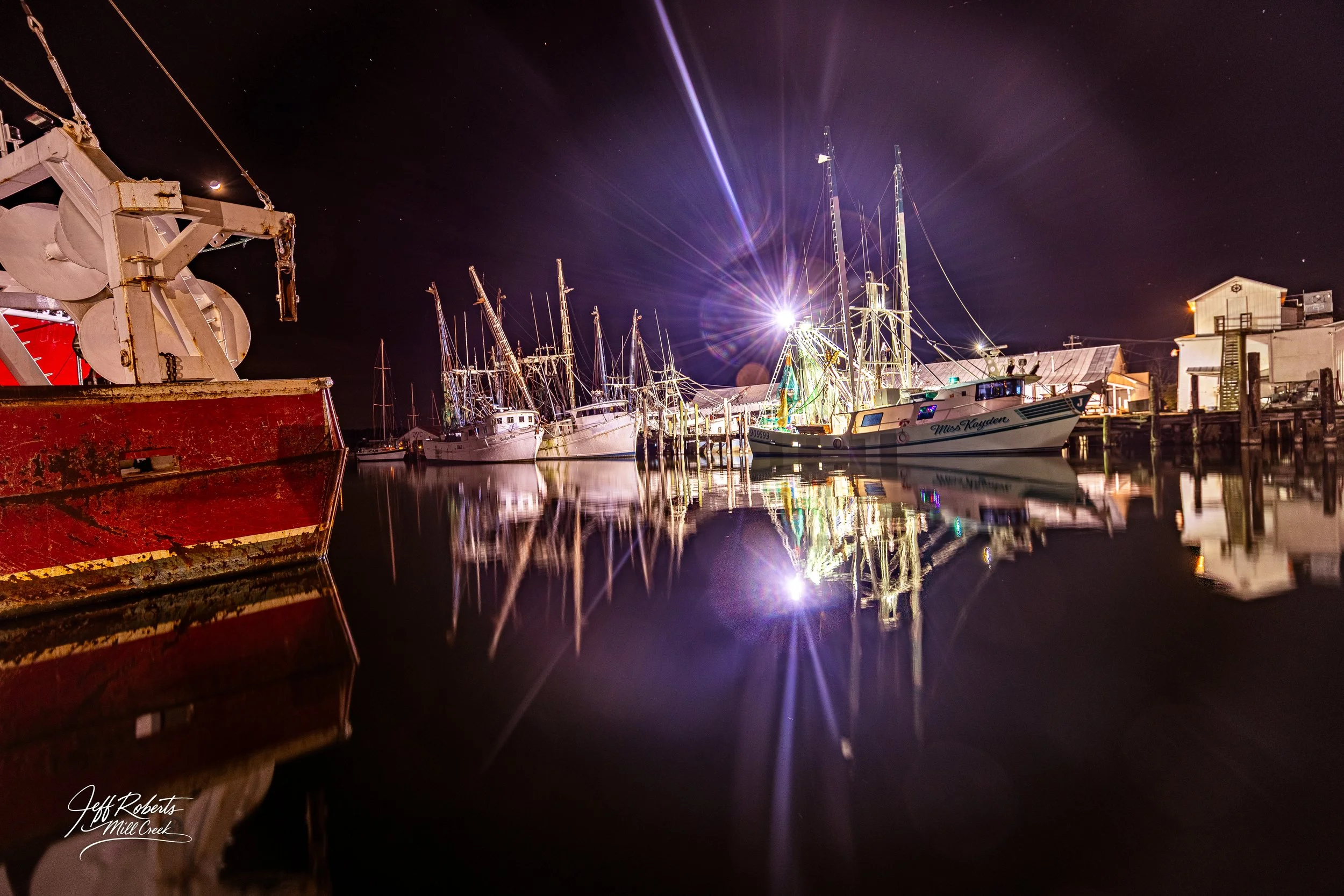 Nighttime scene of a marina with boats moored at docks, bright lights illuminating the area, and reflections on calm water.