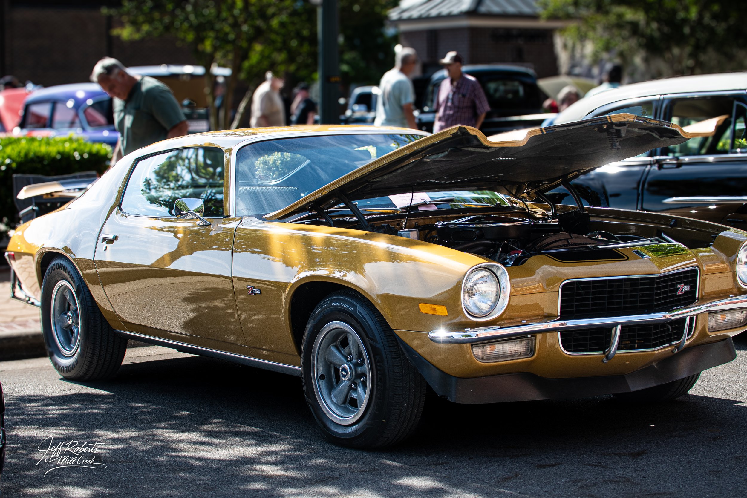 A gold vintage Chevrolet Camaro Z28 muscle car at a car show with its hood open, surrounded by people and other classic cars.