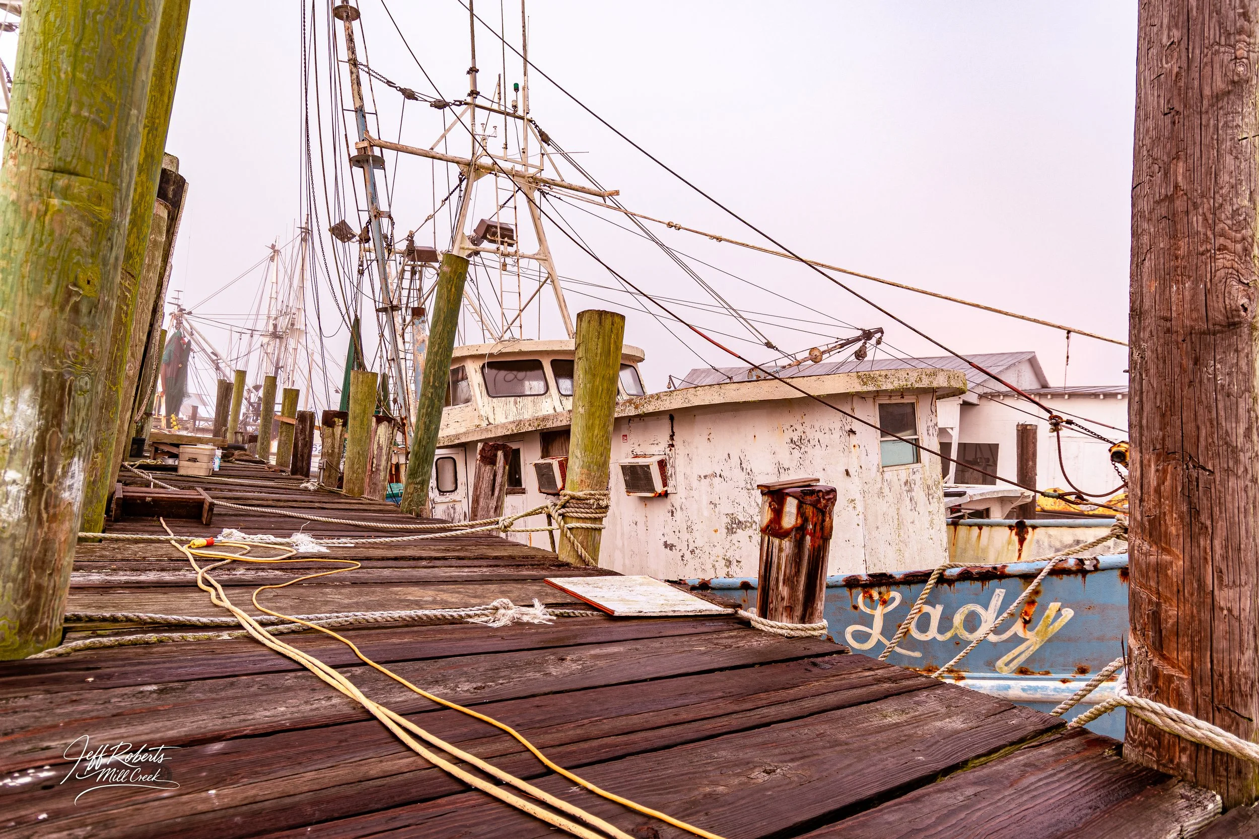 An old, weathered fishing boat tied to a wooden dock with ropes, surrounded by weathered wooden pilings, in a foggy harbor.