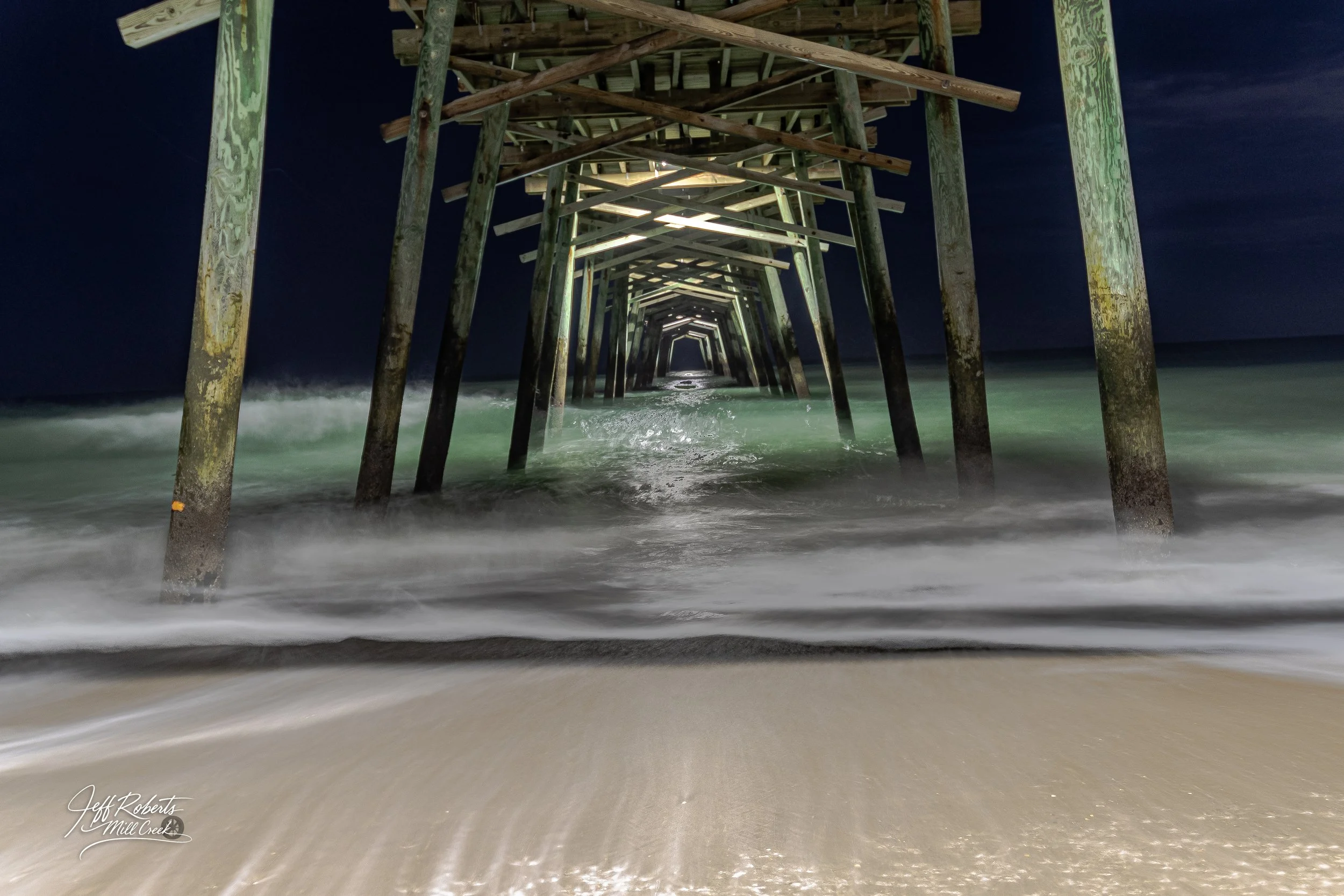 Nighttime view of a wooden pier extending into the ocean with waves crashing underneath, captured from below the pier.