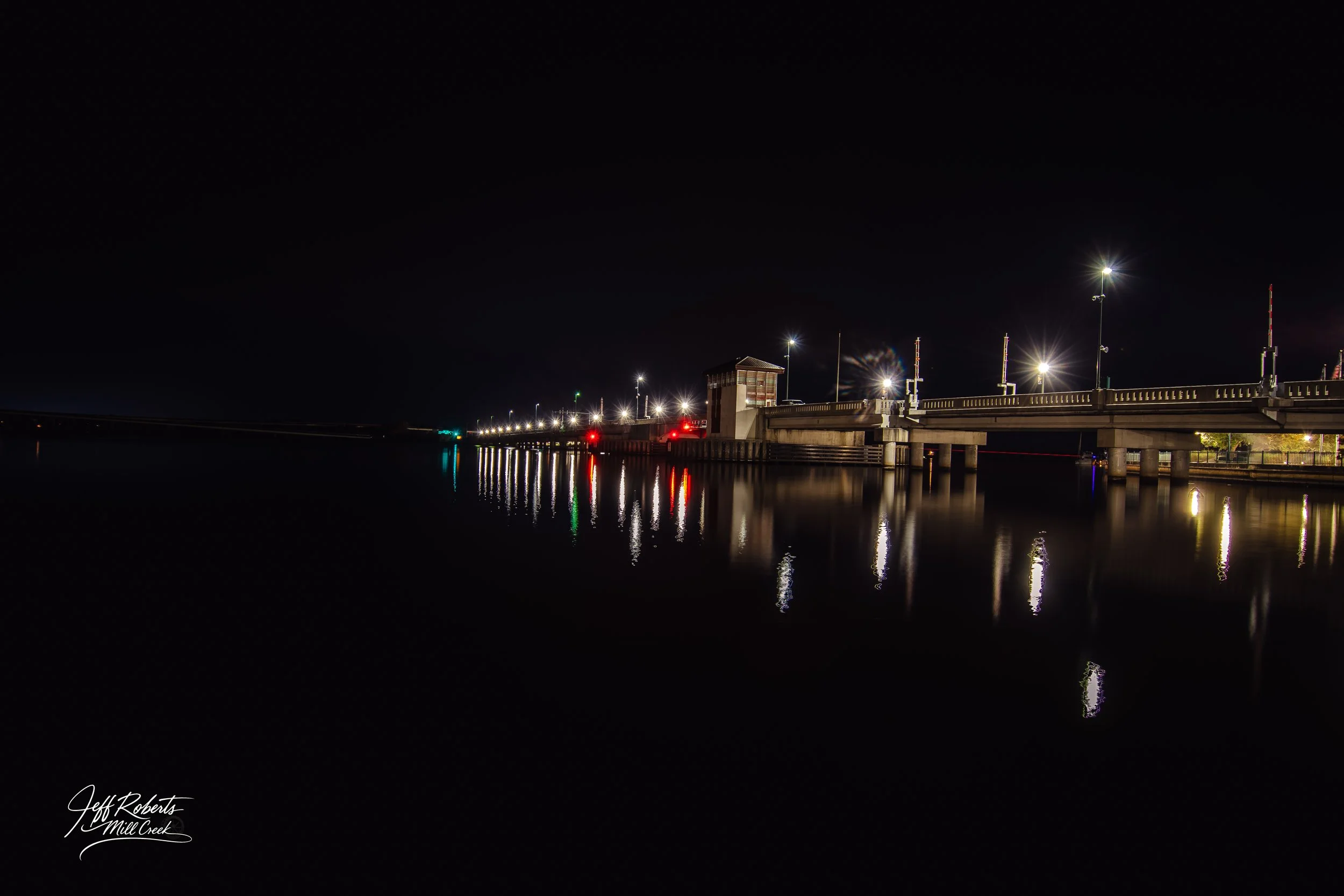 Nighttime image of a bridge with bright streetlights, reflected in calm water below, with a dark sky overhead.