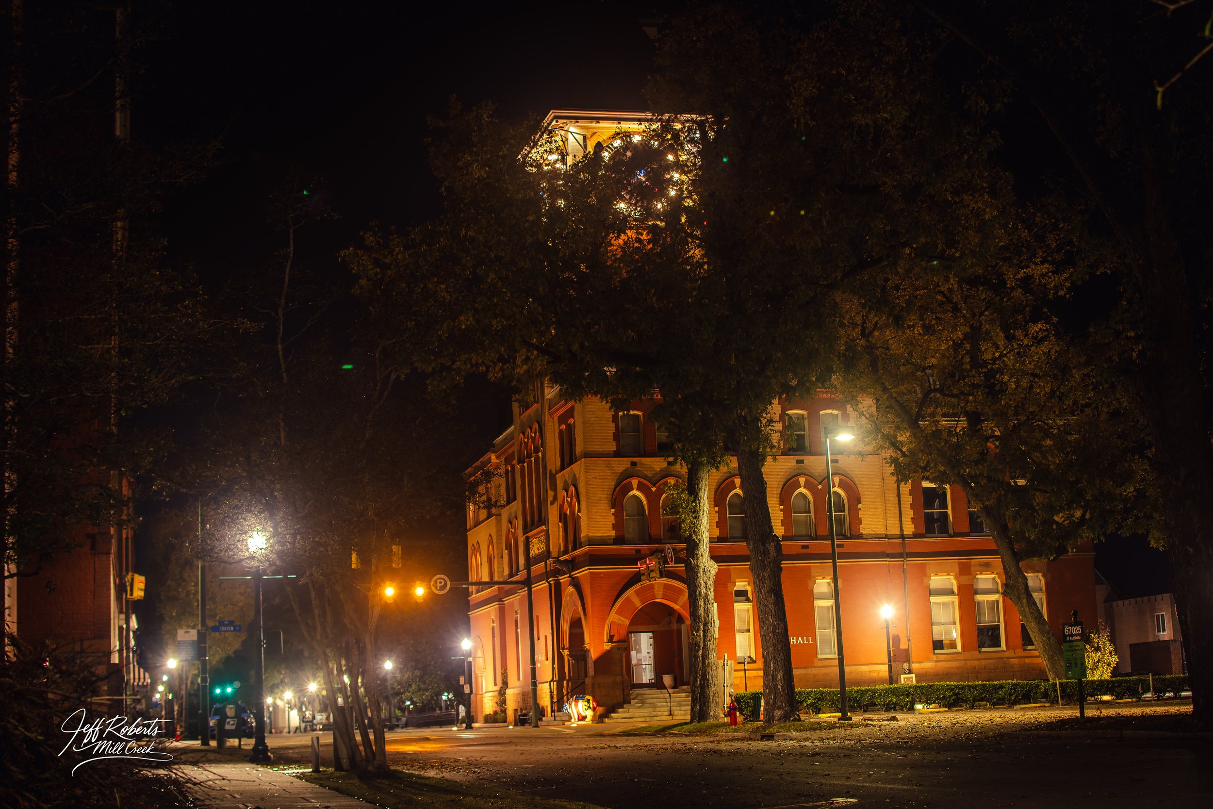 Nighttime street scene featuring a historic brick building illuminated by streetlights and surrounded by trees.