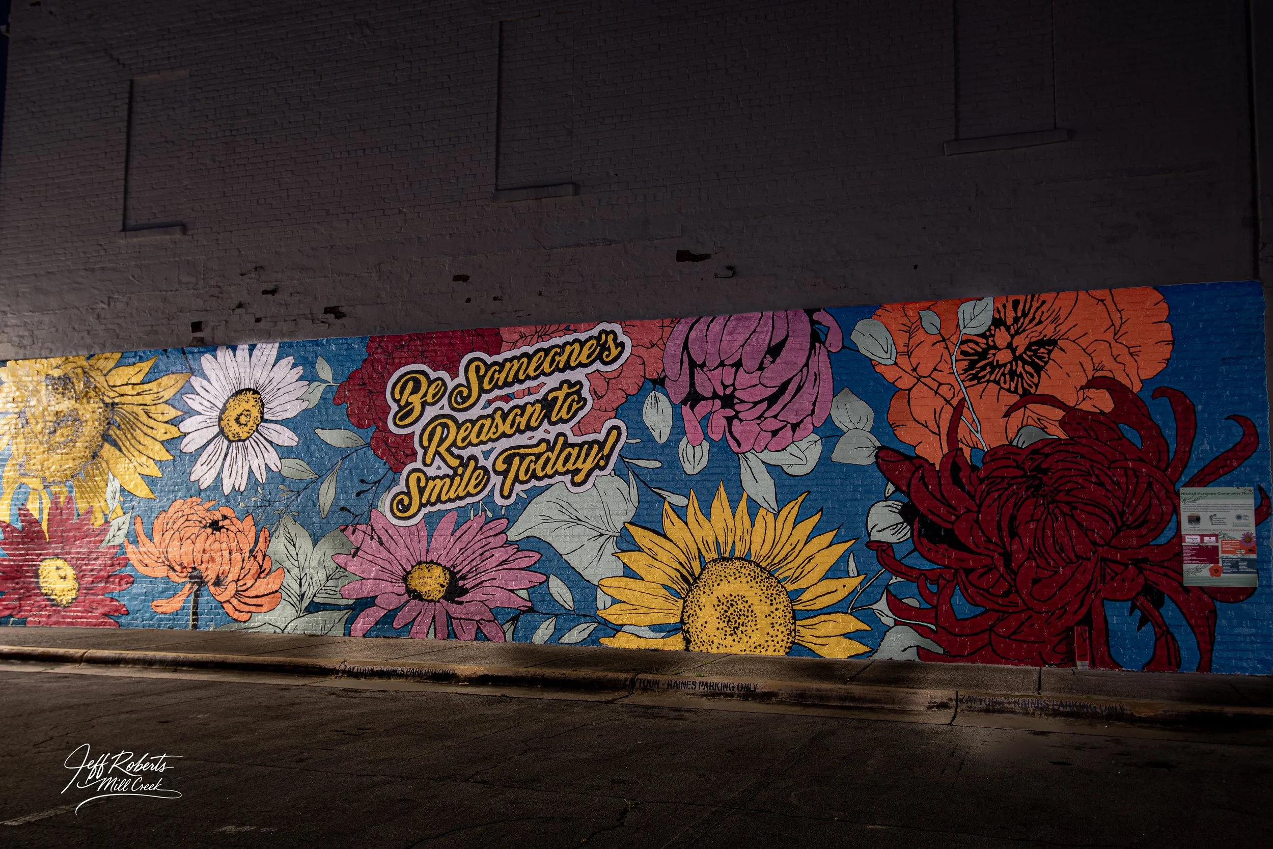 Colorful mural of various flowers, including sunflowers and daisies, with a message that reads, "Be Someone's Reason to Smile Today!" in the center, located under an overpass or in a parking area.